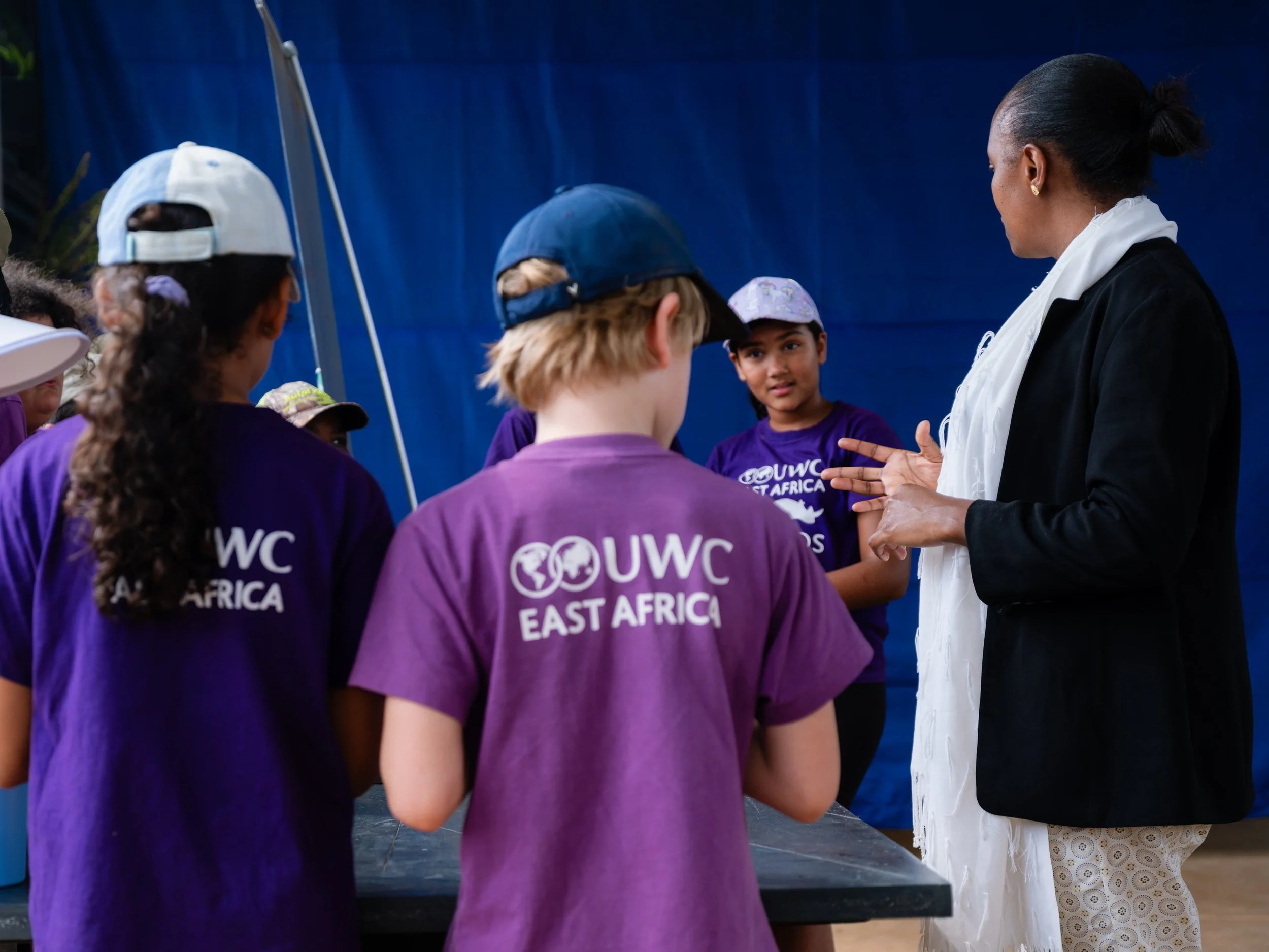 A woman with black hair in a bun speaking to a group of children wearing purple T-shirts with 'UWC East Africa' printed on the back. The children are facing away from the camera and listening attentively.