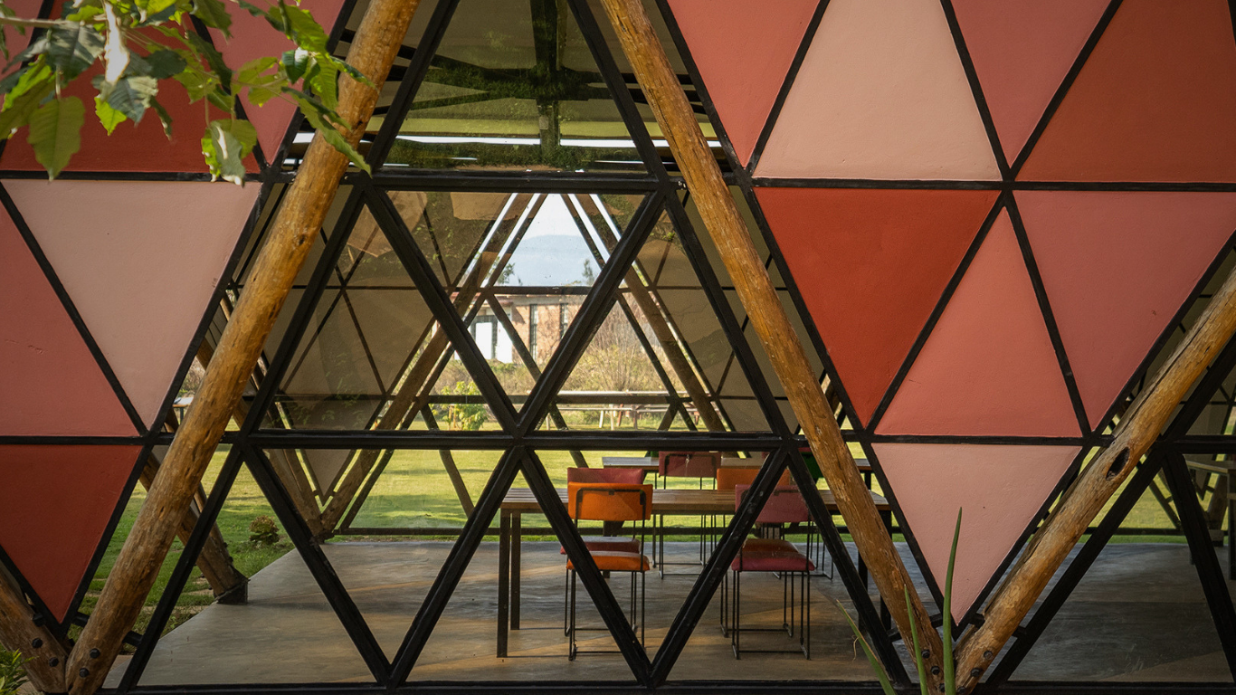 Interior of a geodesic dome with wooden supports, glass walls, and pink triangular panels, featuring a dining table and chairs visible through the structure, with grass and trees outside.