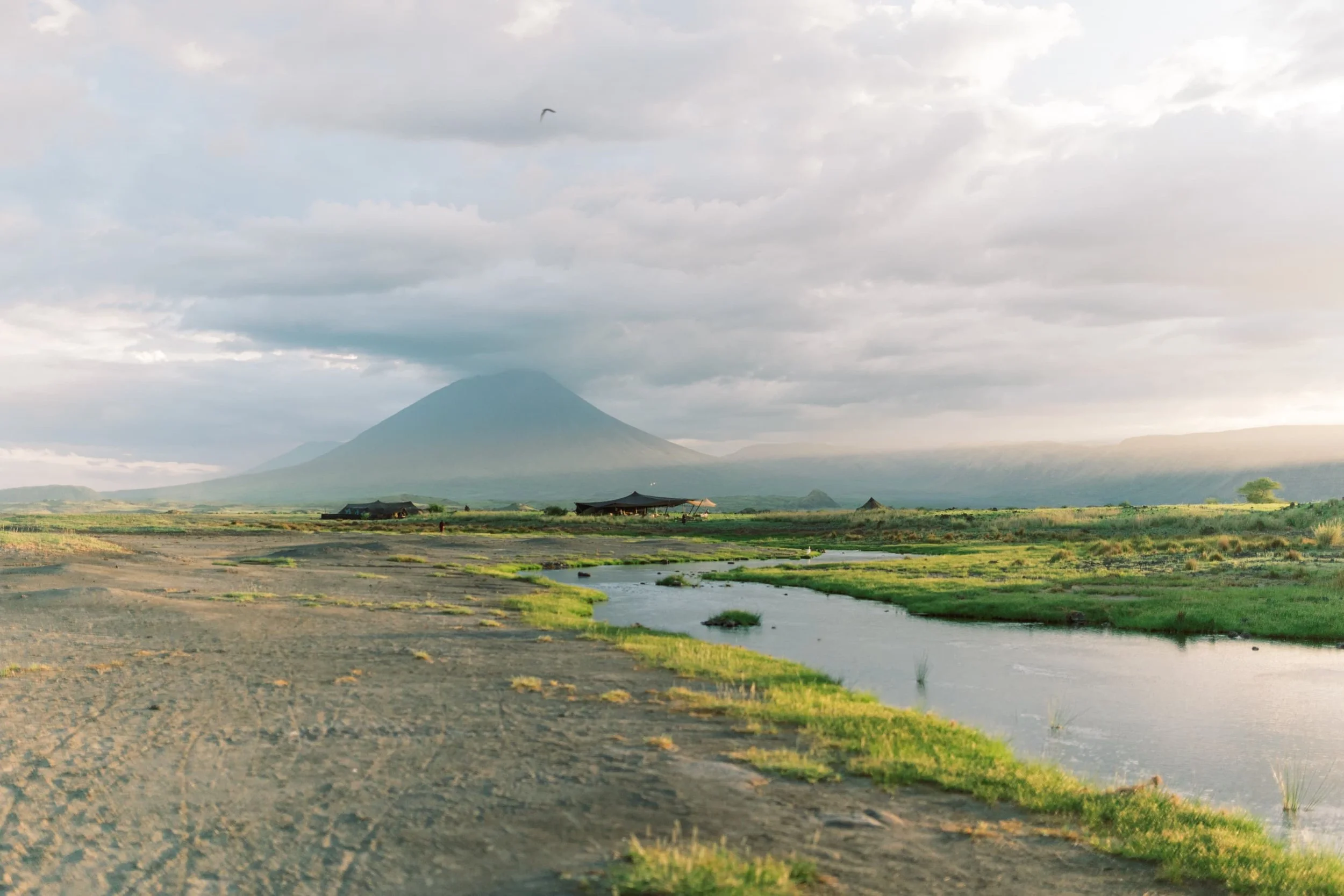 A landscape with a mountain in the distance, cloudy sky, a small river or stream in the foreground, some green grass and a few structures with thatched roofs.