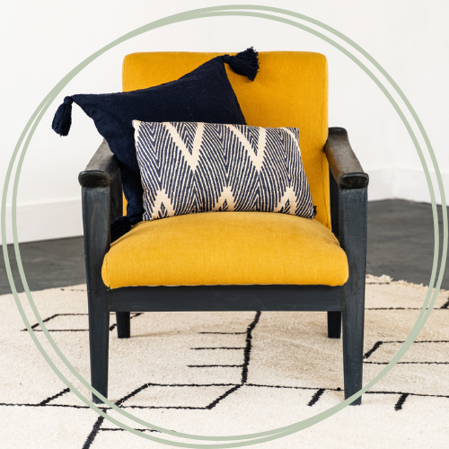 Yellow armchair with black wooden arms and black legs, decorated with a navy blue pillow with tassels and a rectangular patterned pillow, placed on a white rug with black geometric lines.