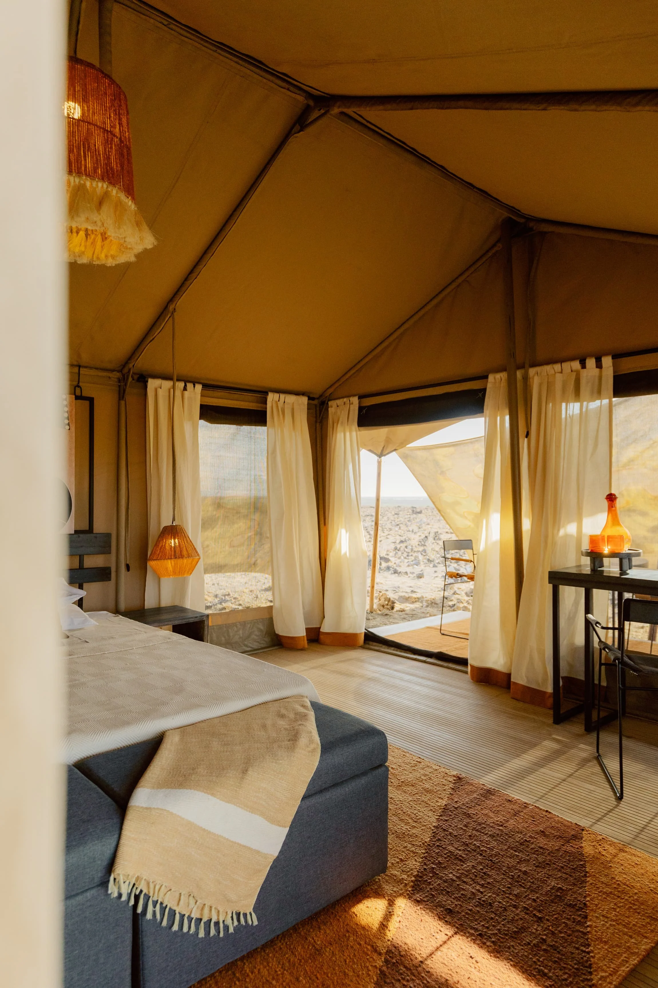 Beach tent interior with a bed, a blue couch with a beige and white throw blanket, a wooden side table, a lamp, curtain panels, and a view of the beach outside.