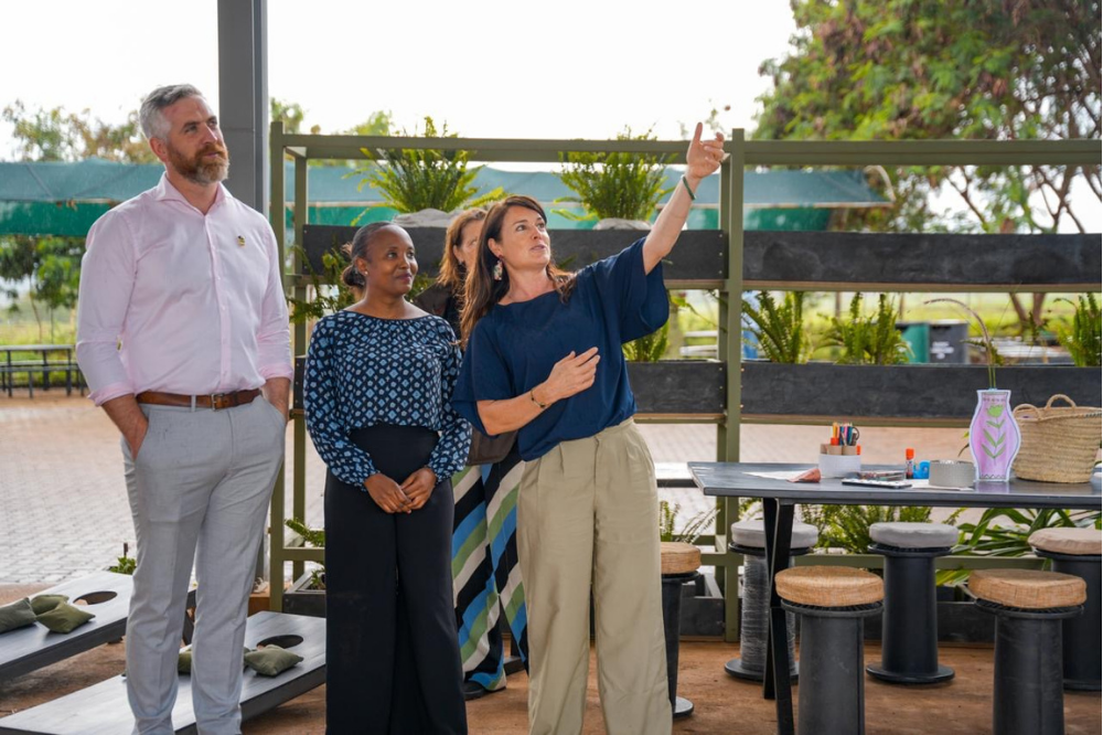 Four people, including a woman pointing upward, standing next to a table with art supplies outside, with a fenced garden area and trees in the background.
