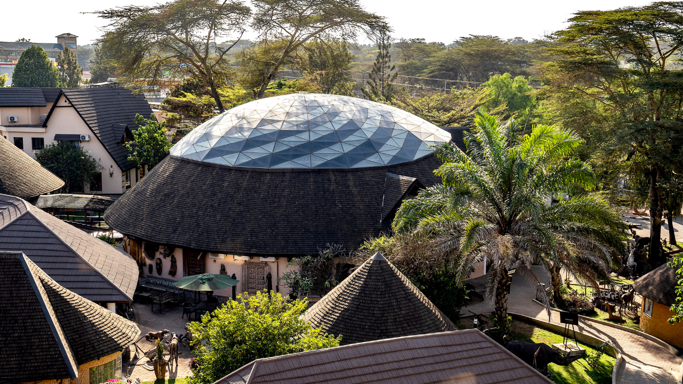 A bird's eye view of a circular building with a geodesic glass dome roof, surrounded by trees, other buildings, and a paved area with outdoor furniture.