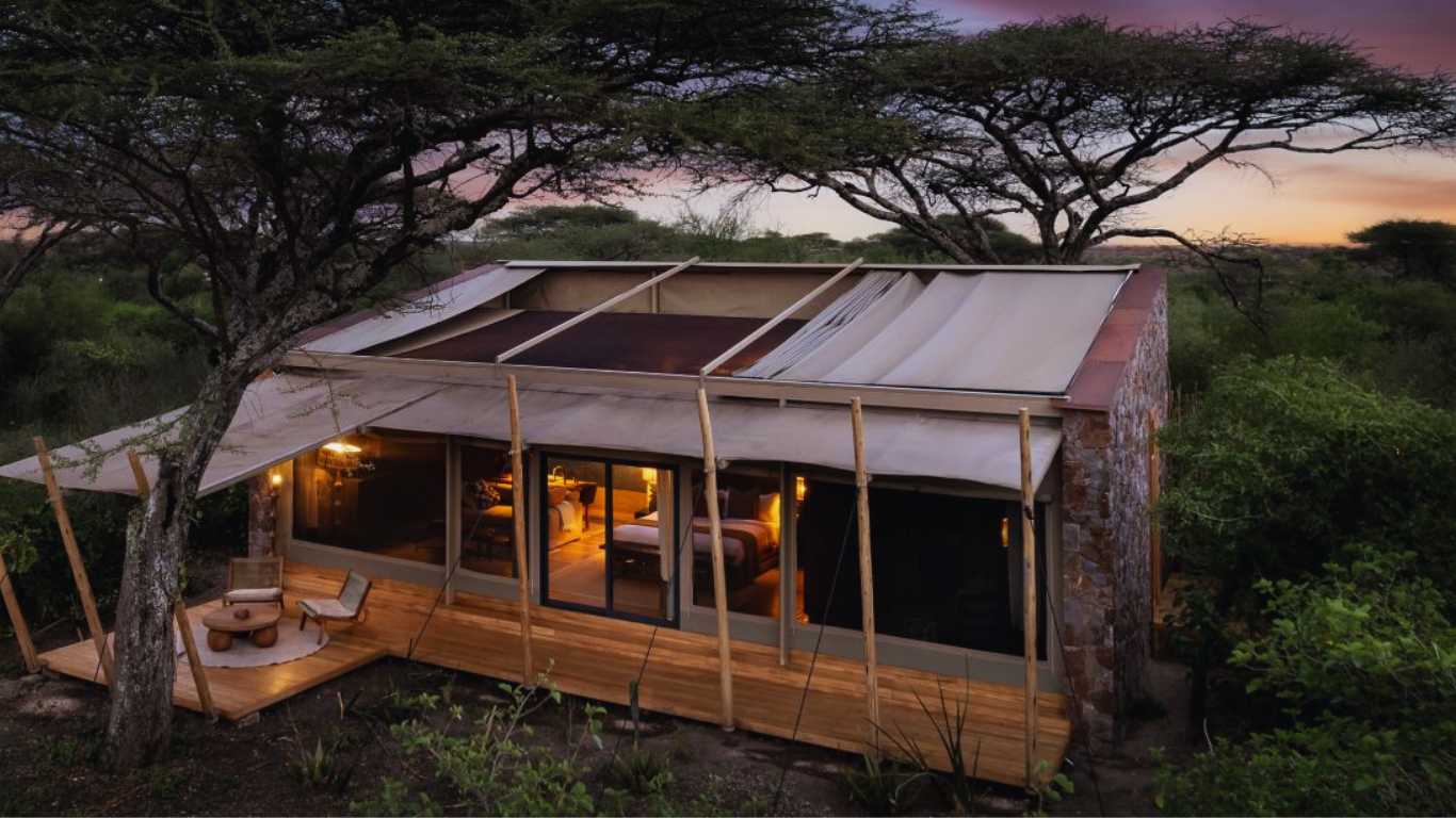 A modern house with large glass windows and a wooden deck, situated among trees in a natural setting during sunset.