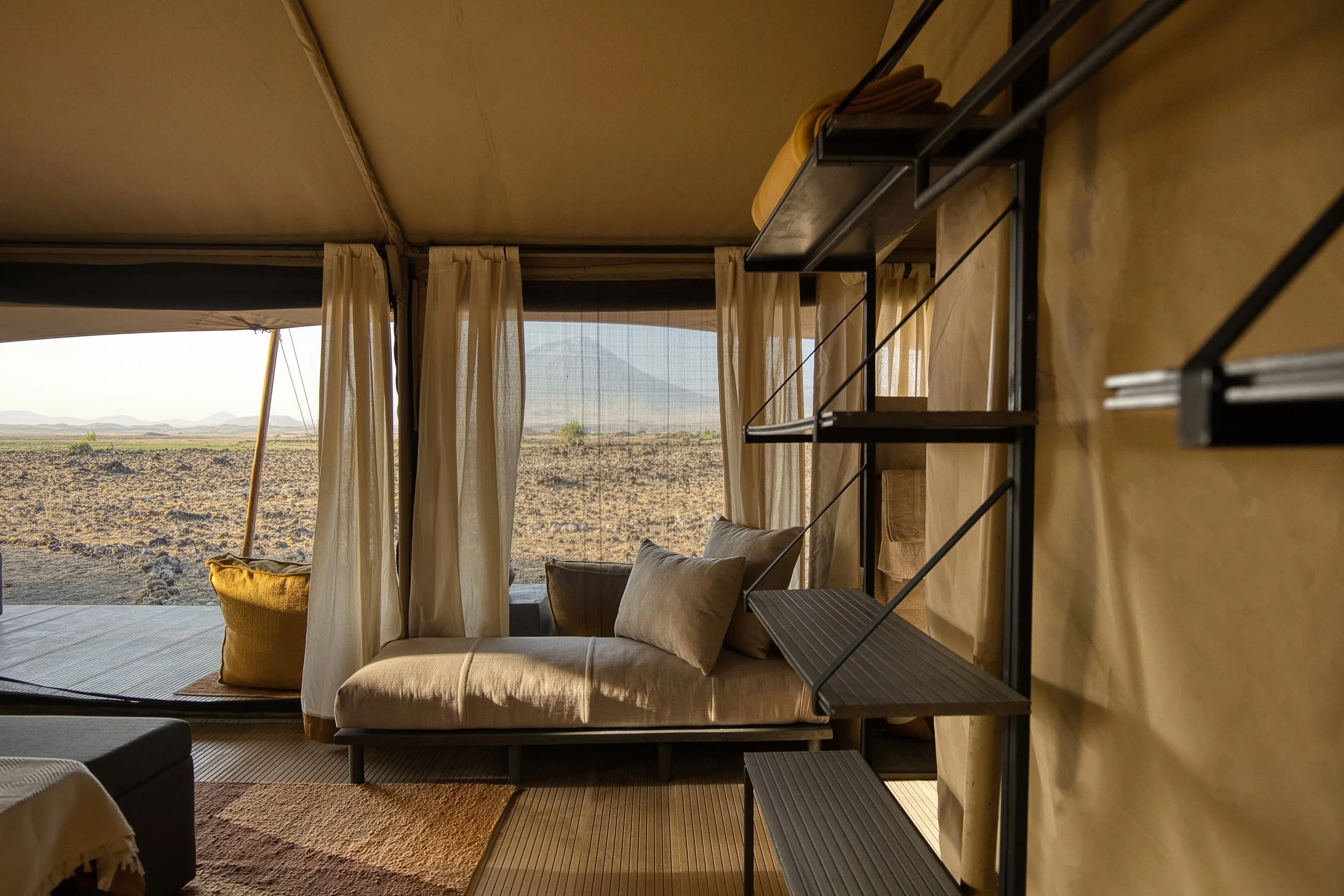 Interior of a cozy room with beige couches, pillows, beige curtains, and a view of a desert landscape with mountains in the distance through an open window.