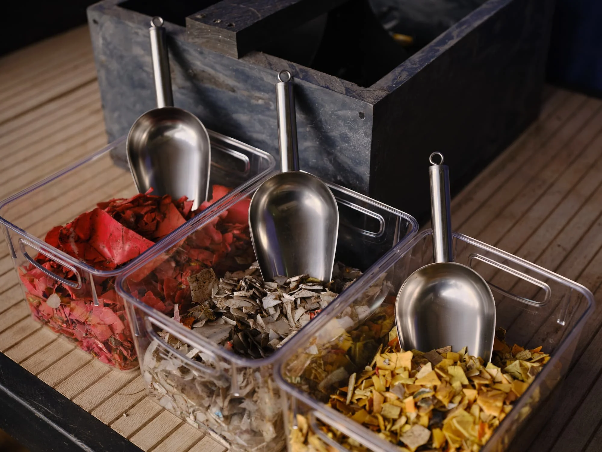 A transparent container with different colored crushed materials and three metal scoops inside, placed on a wooden surface.