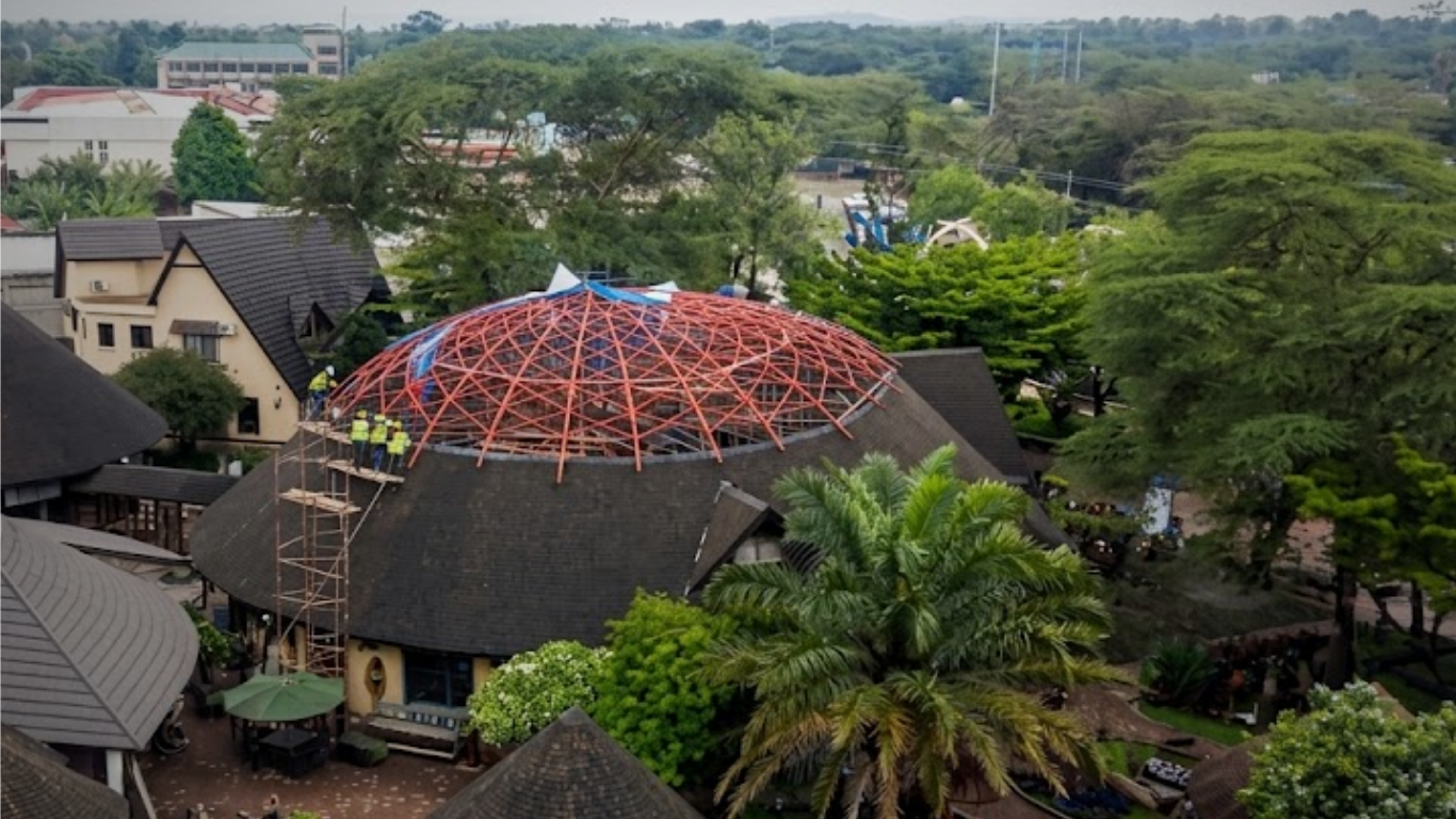 An aerial view of a residential area with a large tree and several houses. There is a dome-shaped roof under construction with a red framework, and workers on scaffolding working on it.