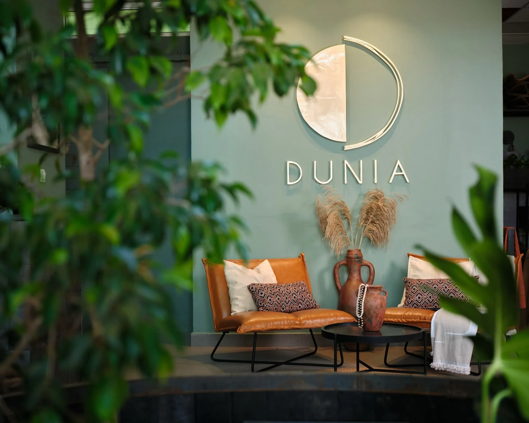 Interior of a furniture store or showroom with a green wall featuring a round mirror and the word 'DUNA' displayed below it. In the foreground, a green leafy plant partially obscures the view. There is a leather sofa decorated with multiple patterned cushions, a round black coffee table, a decorated vase, and a white table with a cloth. The scene evokes a cozy, stylish living space display.