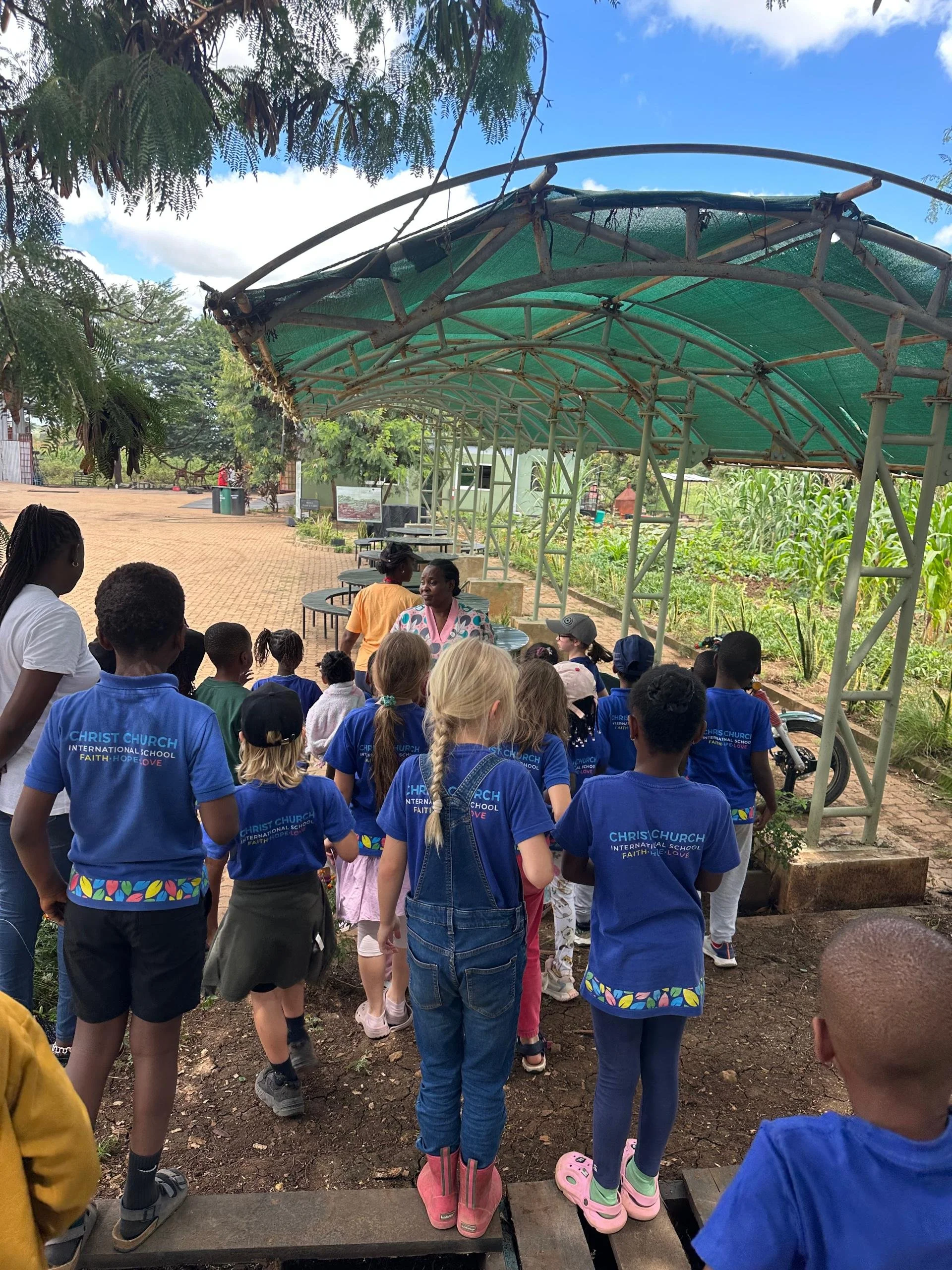A group of children and a few adults gather outside under a green canopy. The children are wearing blue T-shirts with a logo and words, and some are wearing rain boots. A woman in a colorful shirt appears to be speaking to the group. The background s