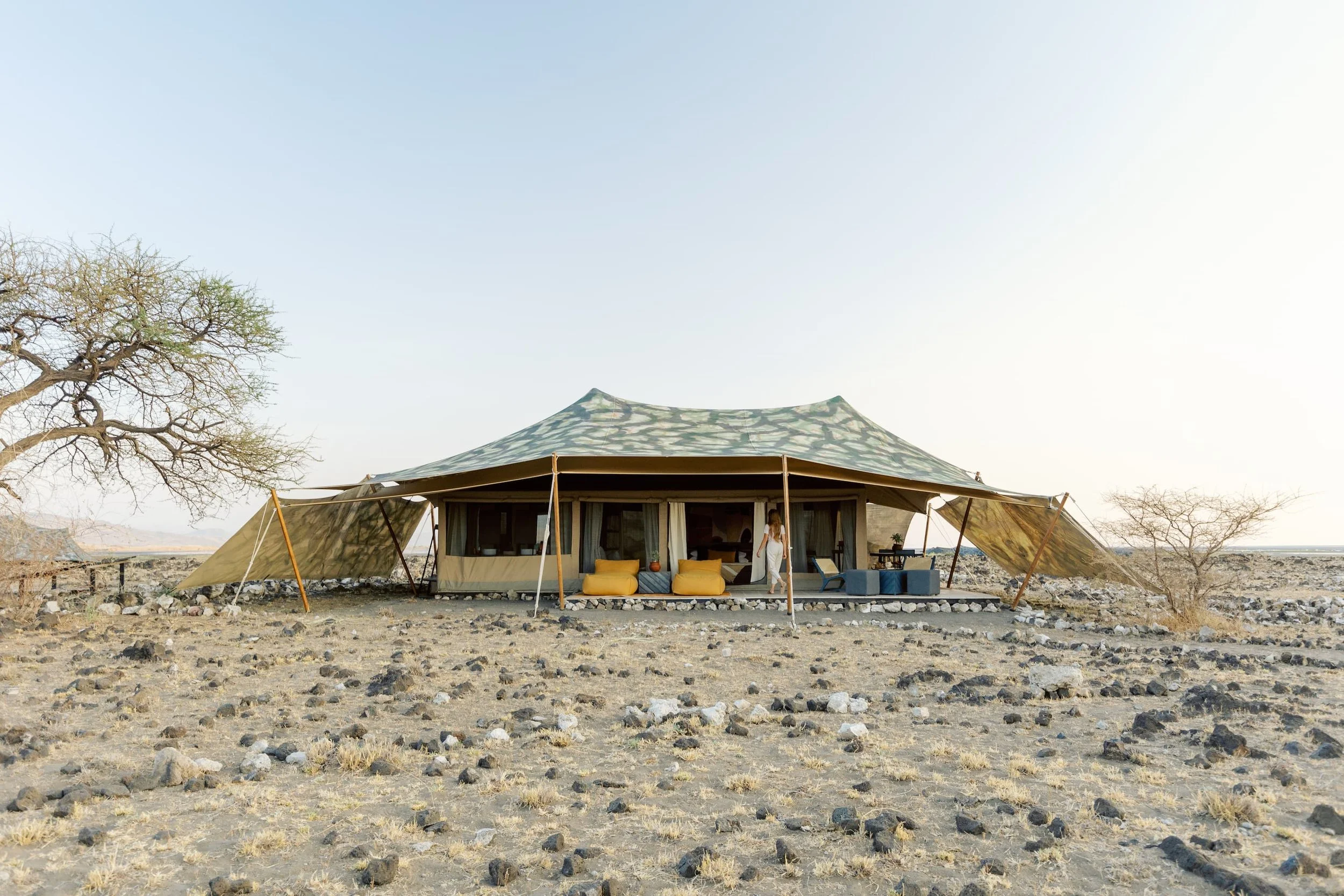 A large canvas tent set up in a dry, rocky desert landscape with sparse trees, furnished with colorful cushions and outdoor furniture, under a clear sky.