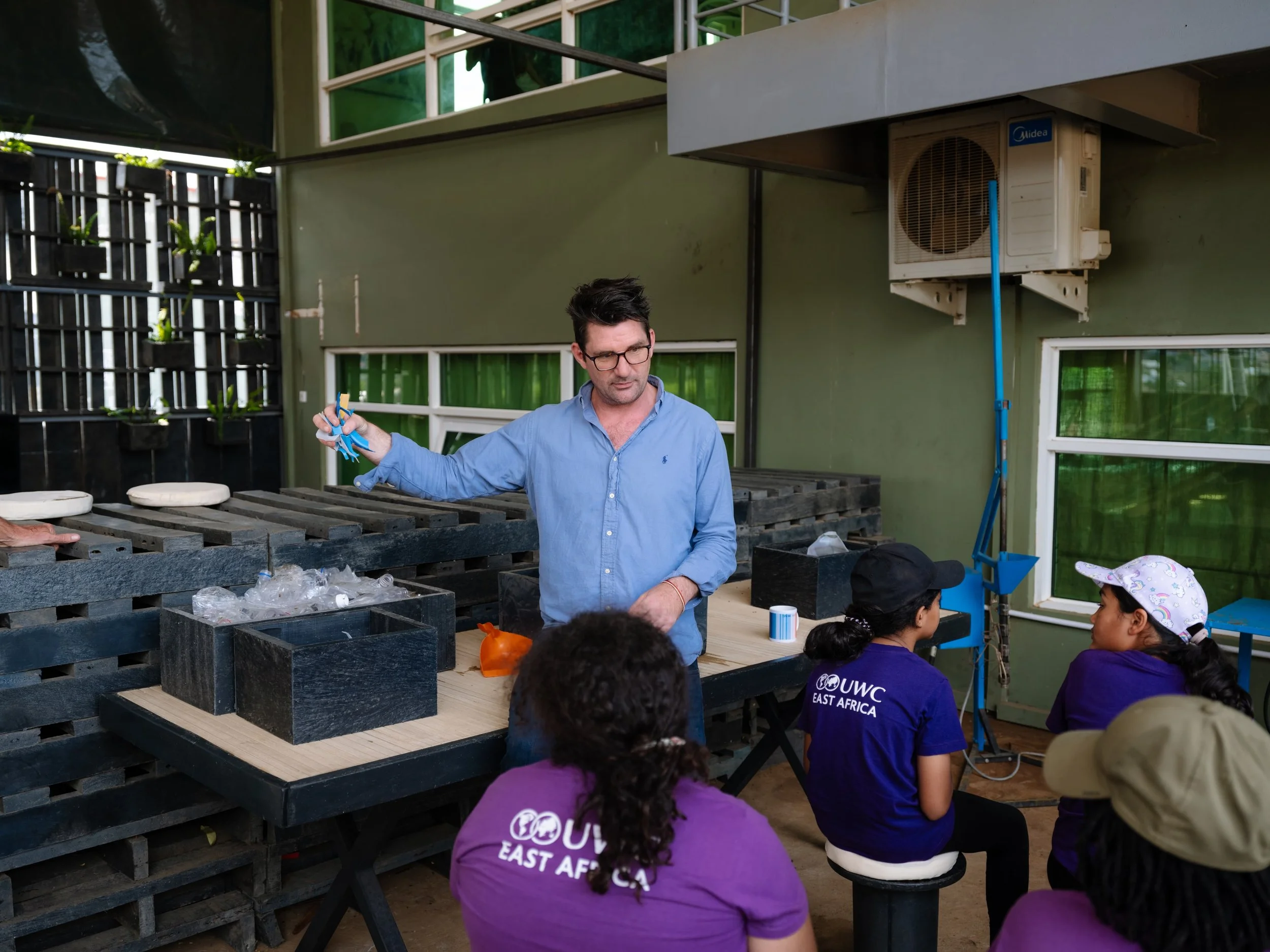 A man with glasses and a blue shirt holding scissors in an outdoor classroom with children wearing purple shirts, some with hats, sitting on benches, and a green building with windows and an air conditioning unit in the background.