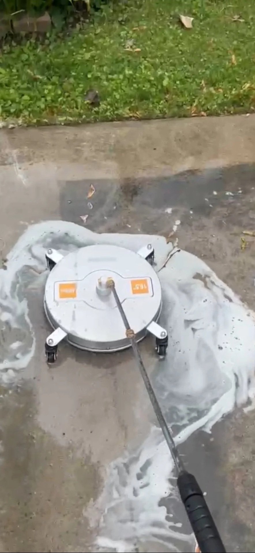 A person using a pressure washer to clean the concrete sidewalk, with a grassy area and fallen leaves in the background.