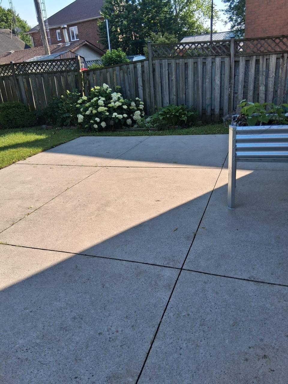 A backyard patio with concrete tiles, a small grassy area, flowering bushes, wooden fence, surrounding houses, and a metal bench.
