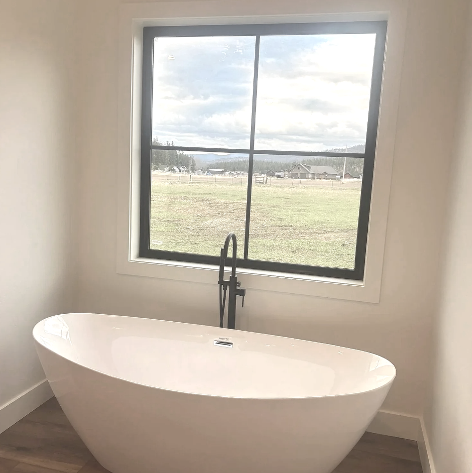 Modern white bathtub with black faucet, positioned in front of a large window showing an outdoor landscape with fields, trees, and houses.