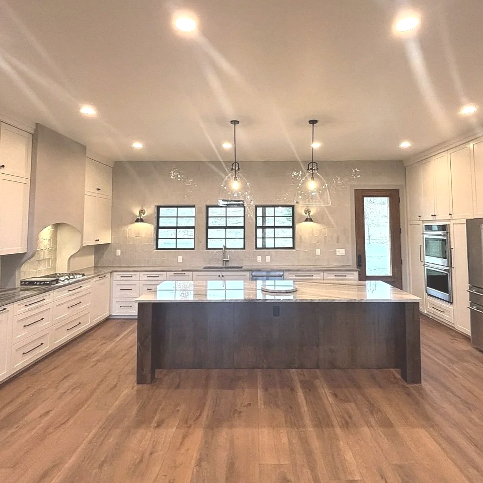 Modern kitchen with a large island, white cabinets, three windows, pendant lighting, and a wooden door.