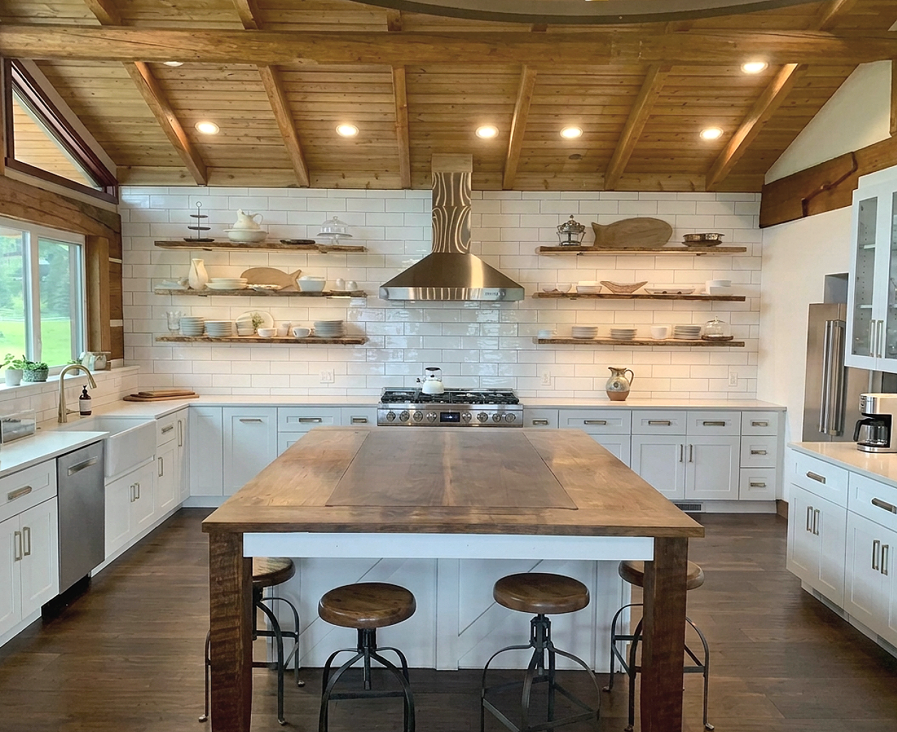Modern kitchen with white cabinets, wooden open shelves, and a large kitchen island with stools. There is a stainless steel range hood above a stove, and the ceiling is made of wood with recessed lighting. Large windows provide natural light, and the walls are tiled with white subway tiles.