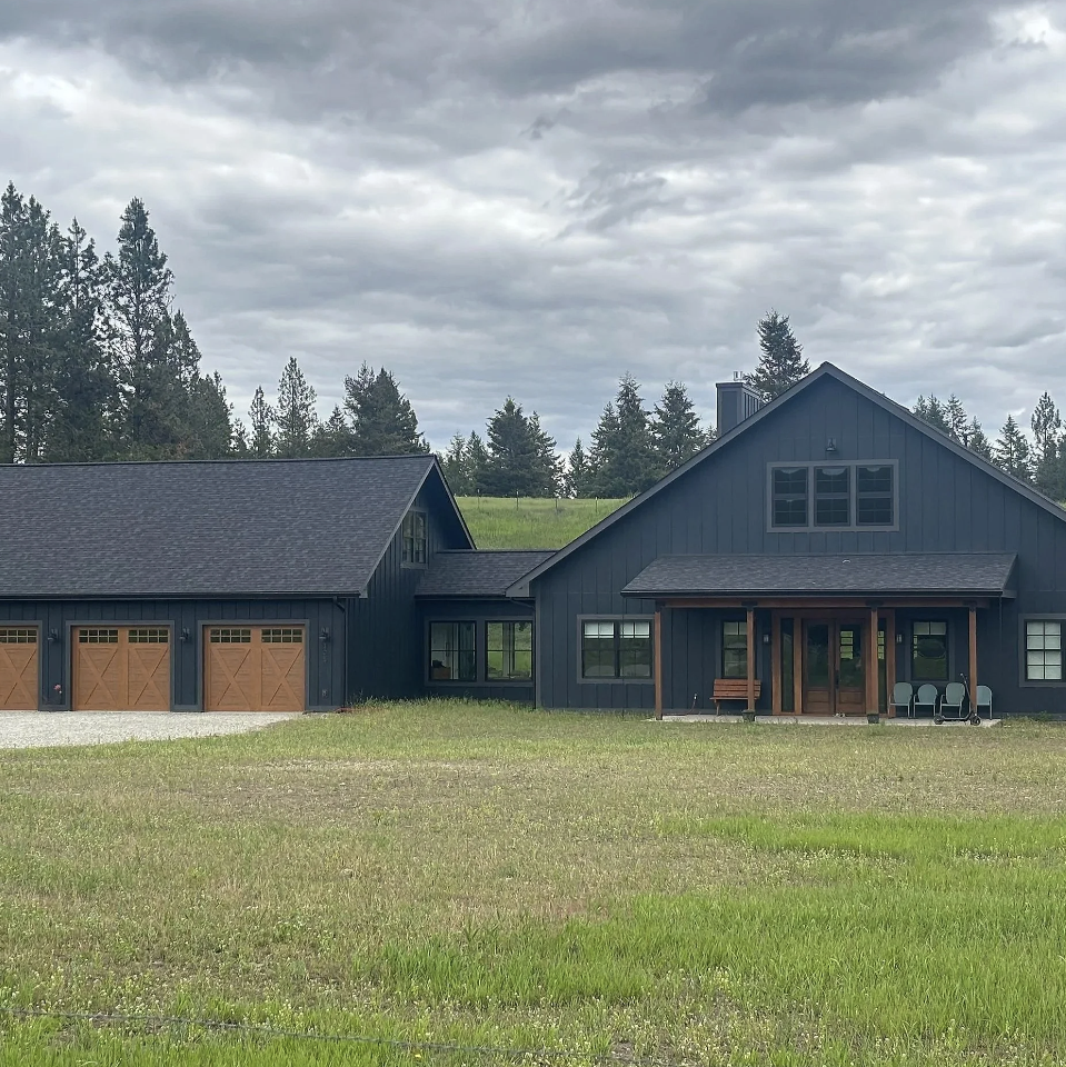 A modern dark blue house with a three-car garage and wooden accents, set on a grassy field under a cloudy sky with trees in the background.