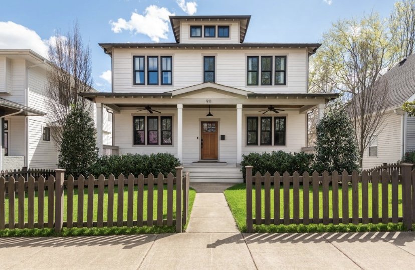 Front view of a modern, white, three-story house with a brown wooden front door, black framed windows, and a small porch with steps. There is a brown picket fence and a green lawn in front.