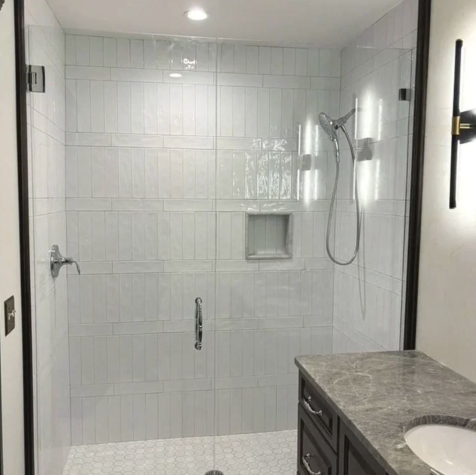 Bathroom shower with glass door, white tile walls, showerhead, built-in niche, and gray marble countertop vanity.