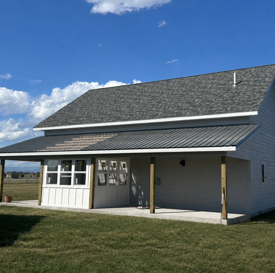 A two-story house with a metal roof, white siding, a covered porch with support beams, and large windows. The house is situated in a grassy field with a blue sky and some clouds.