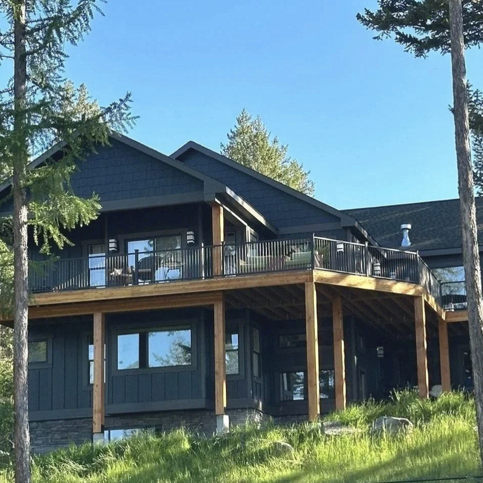 A modern two-story house with outdoor balcony, black siding, surrounded by trees, with a bright blue sky in the background.