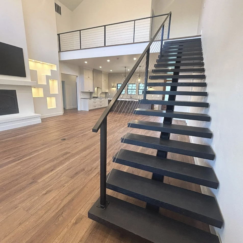 Empty modern living room with black open-riser staircase, white walls, hardwood floors, and kitchen in the background.