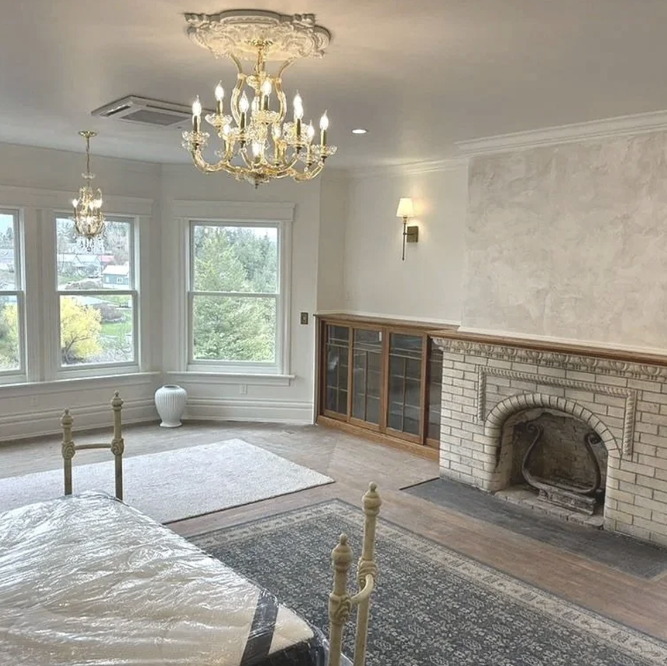 Living room with large bay windows, a chandelier, a fireplace with a brick surround, wall sconces, and a built-in wooden cabinet, with a bed partly visible in the foreground.