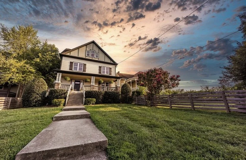A large, two-story house with a front porch, surrounded by a well-maintained lawn, bushes, and trees, under a colorful, partly cloudy sunset sky.