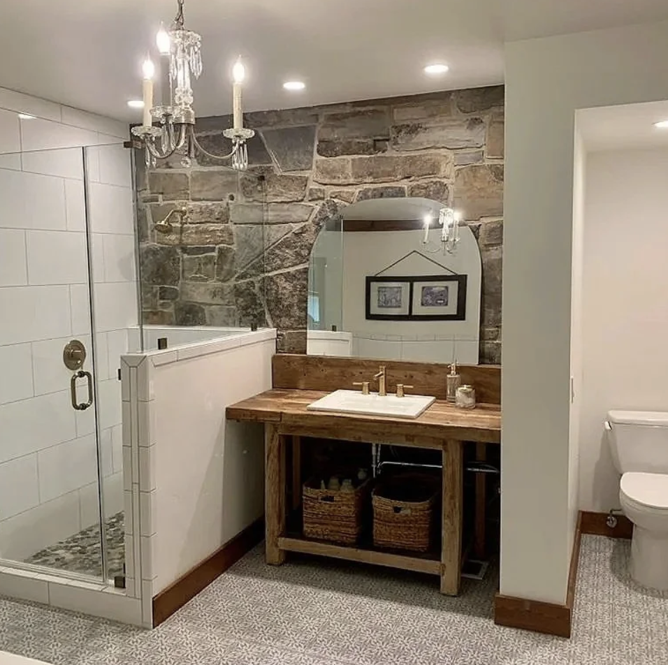 A bathroom with a glass shower, a wooden vanity with a sink, a large mirror, a stone wall, and a chandelier.