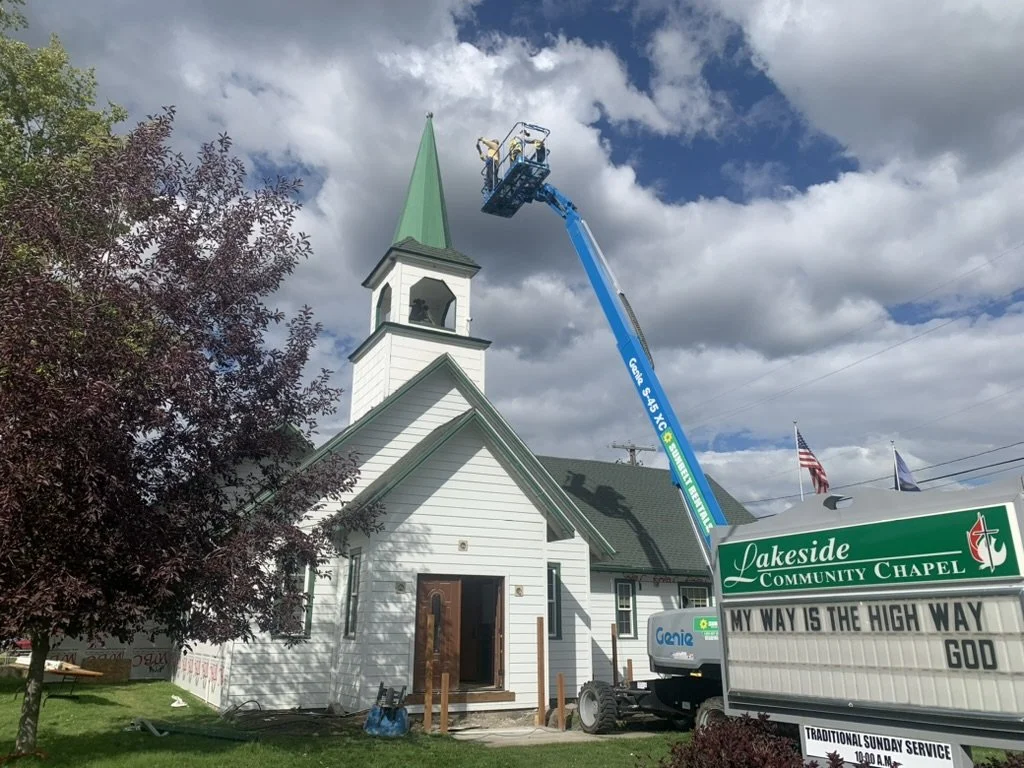 A church with a steeple undergoing maintenance using a cherry picker. The church is white with green roof accents, and there is a sign that reads "Lakeside Community Chapel" with a biblical verse. There are trees and flags nearby, and the sky is partly cloudy.