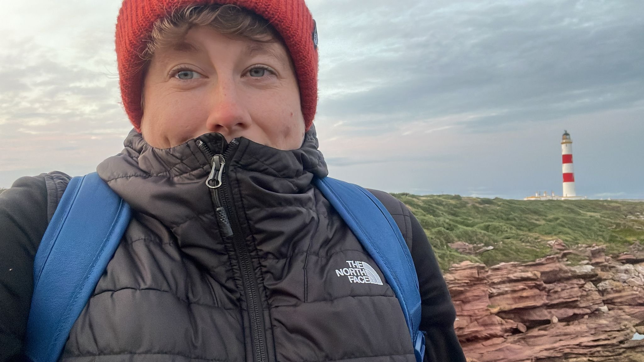 A person with a red knit hat, black outdoor jacket, and blue backpack outdoors with a lighthouse in the distance on a cloudy day.