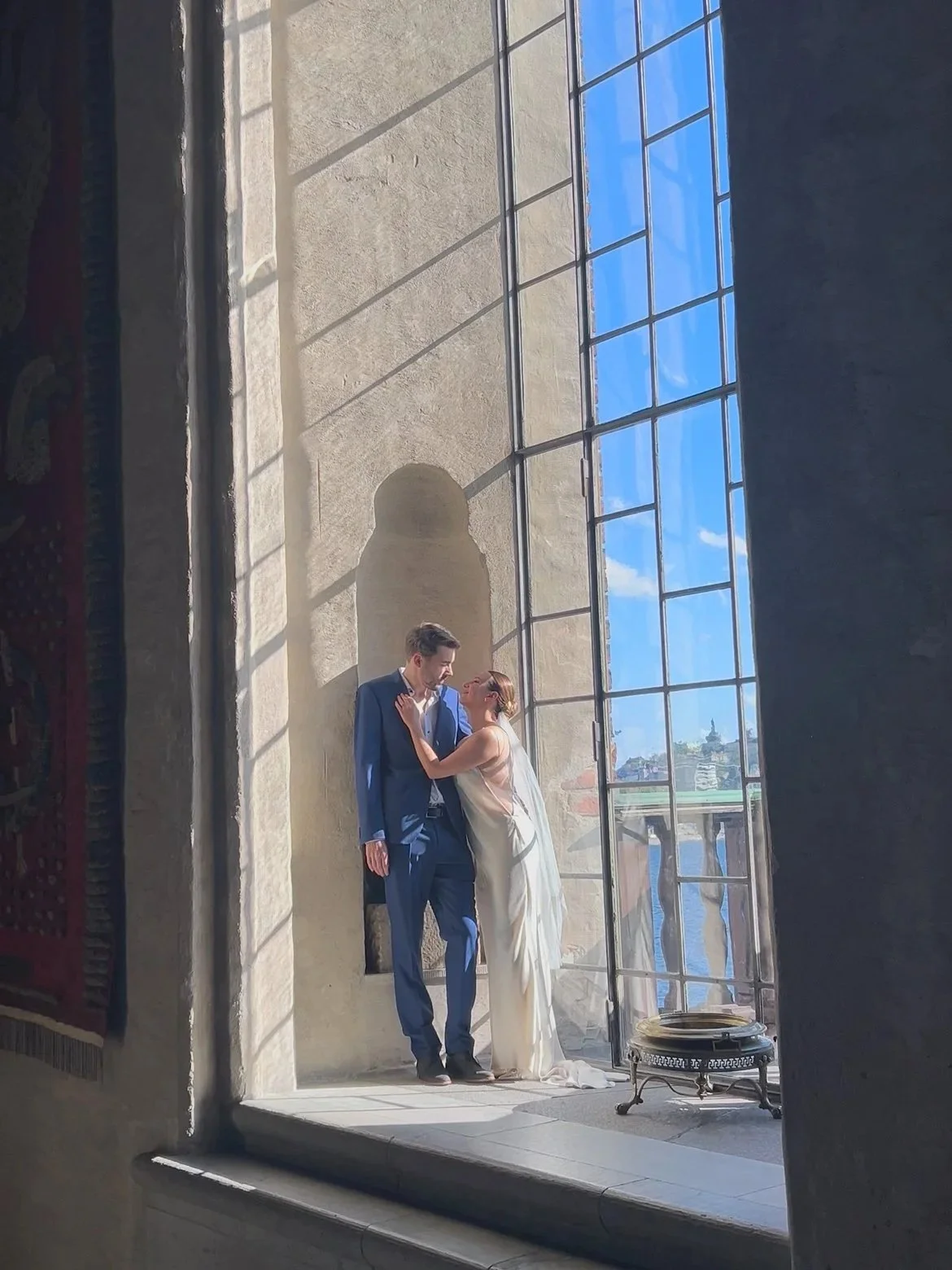 A bride and groom stand inside a building near a large window with a view of the city and river outside. The bride is wearing a white dress, and the groom is in a blue suit. The sunlight streams in, casting shadows on the wall behind them.