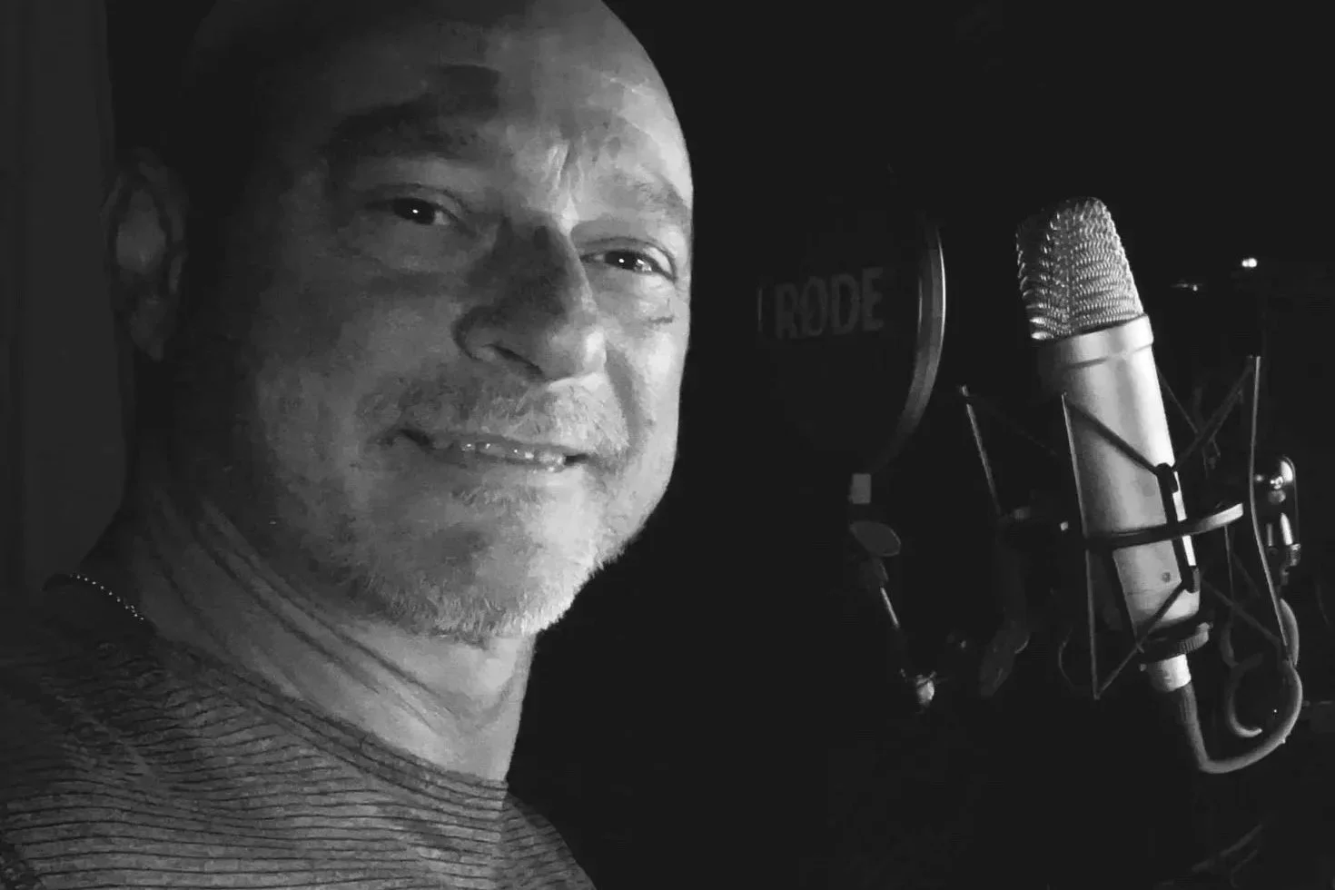 A man in a recording studio takes a selfie with a large microphone in the background.