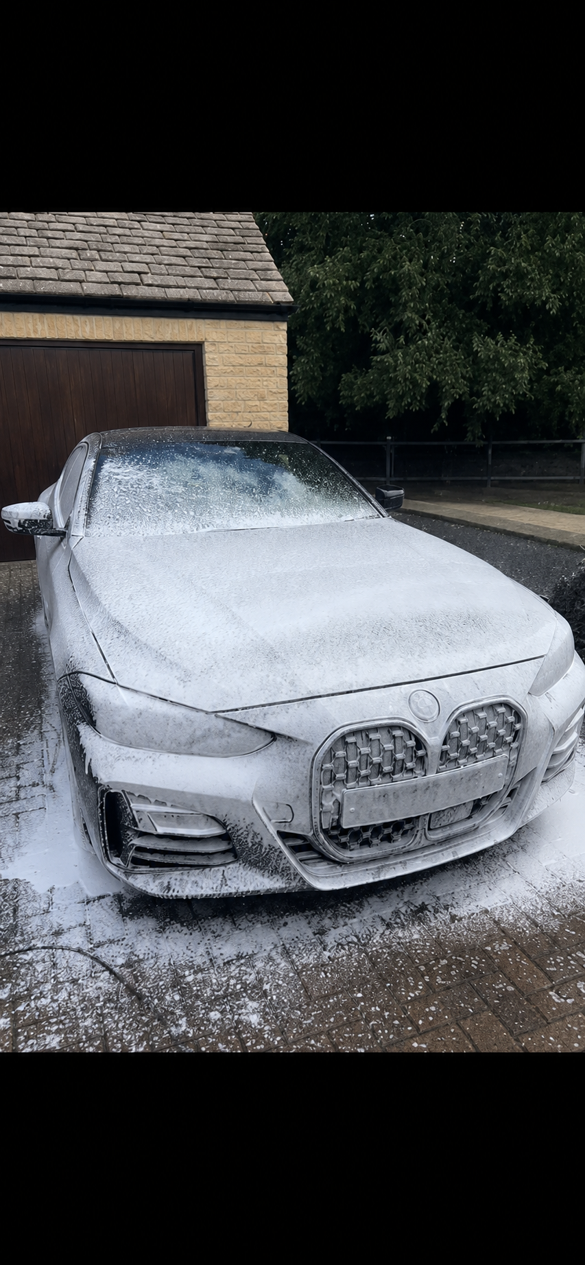 A white car being washed with foam, parked on a driveway near a garage with a brown door, surrounded by trees and greenery.