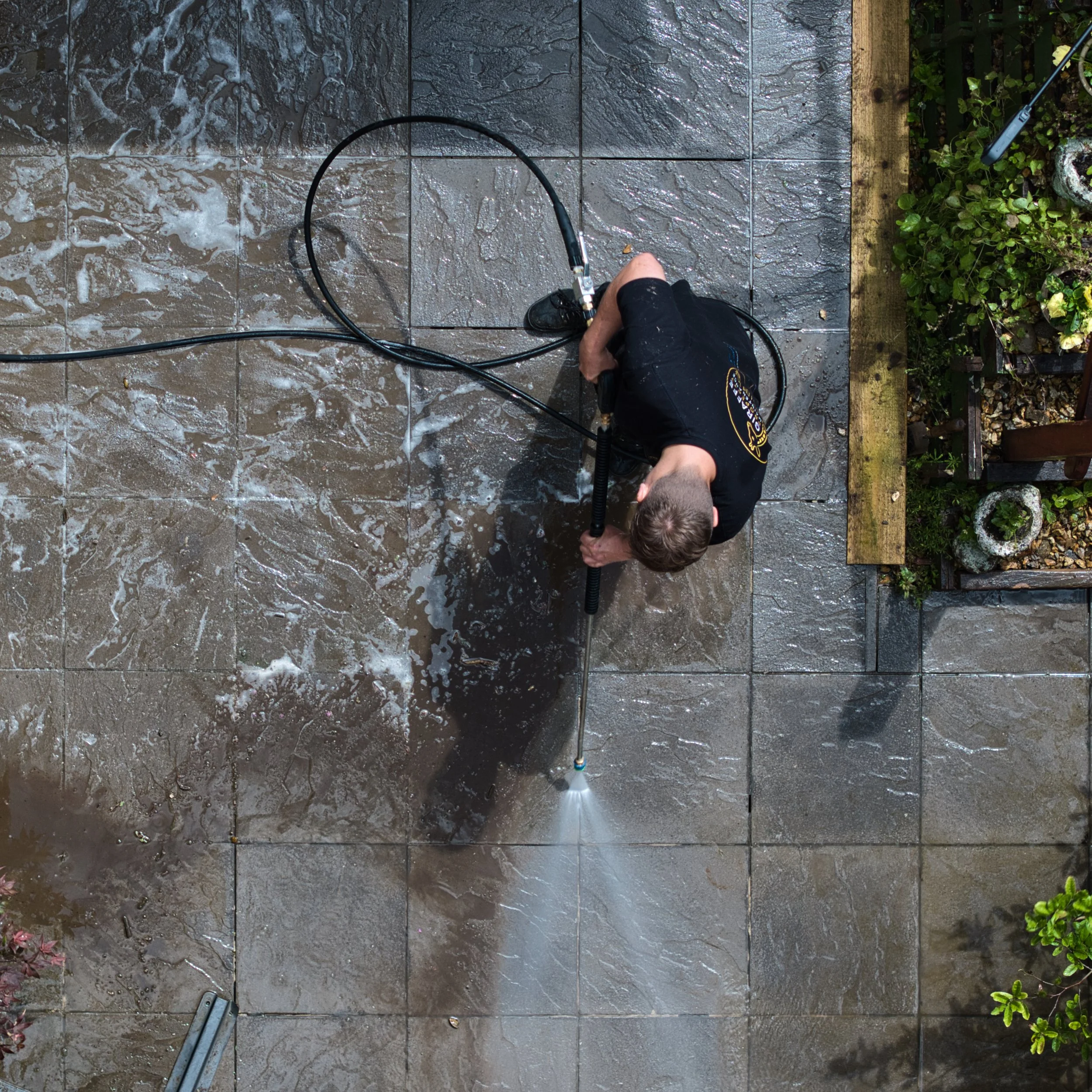 A person using a pressure washer to clean a wet, tiled patio with soap suds, near a wooden border and some green foliage.