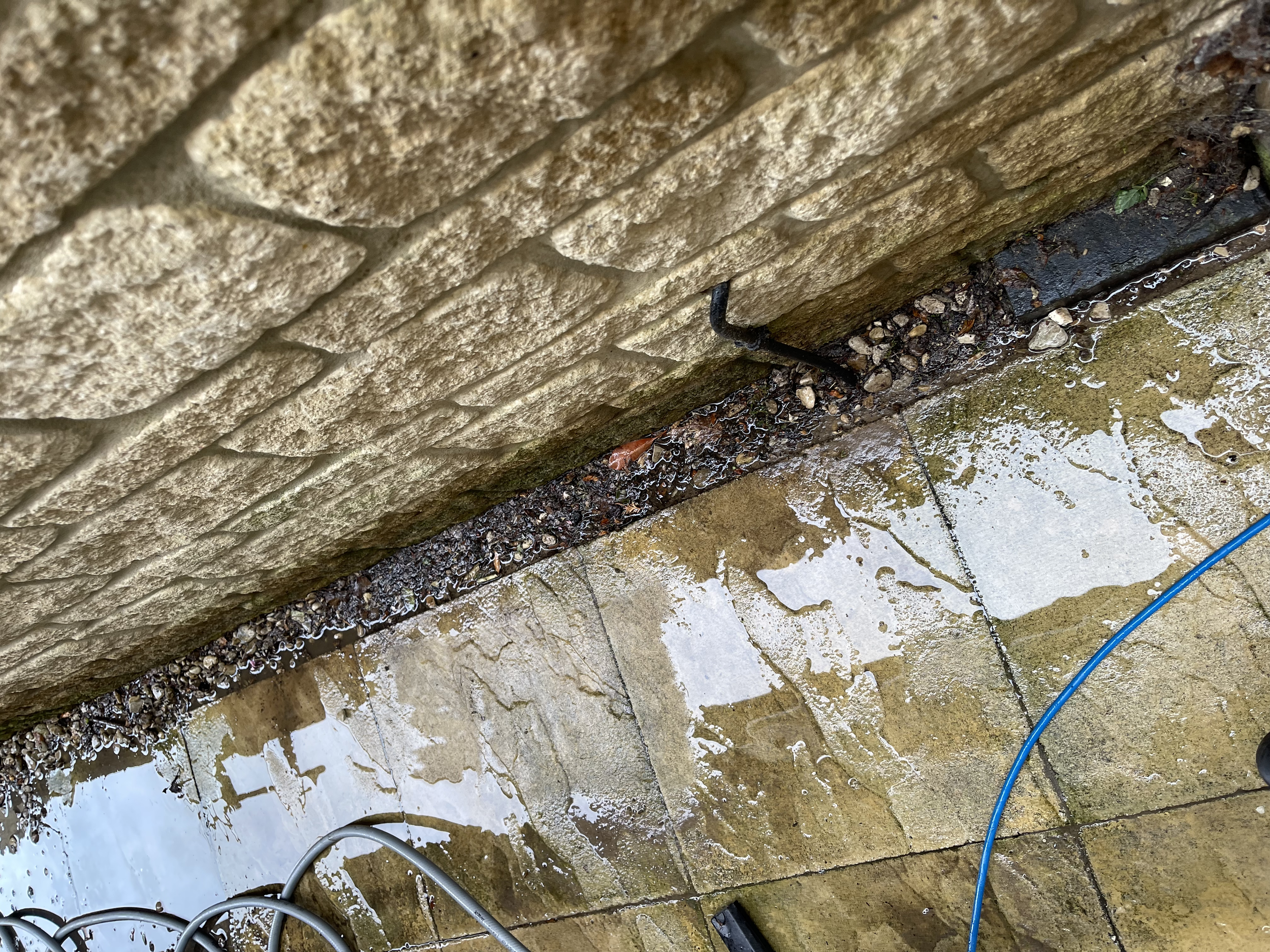 Close-up of a brick wall with black metal hooks and pipes, wet with water, on a stone or concrete ground with scattered debris and a blue hose.