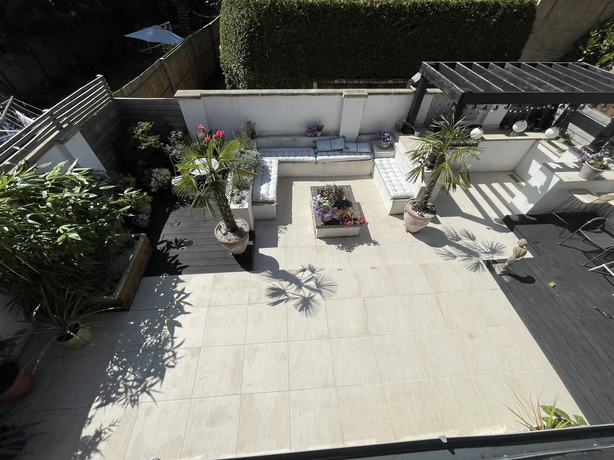 A view of an outdoor patio area with white tiled flooring, potted plants, and an L-shaped white cushioned seating area. There are various potted plants and flowers, a small table with flowers, and some patio furniture including an umbrella in the background.