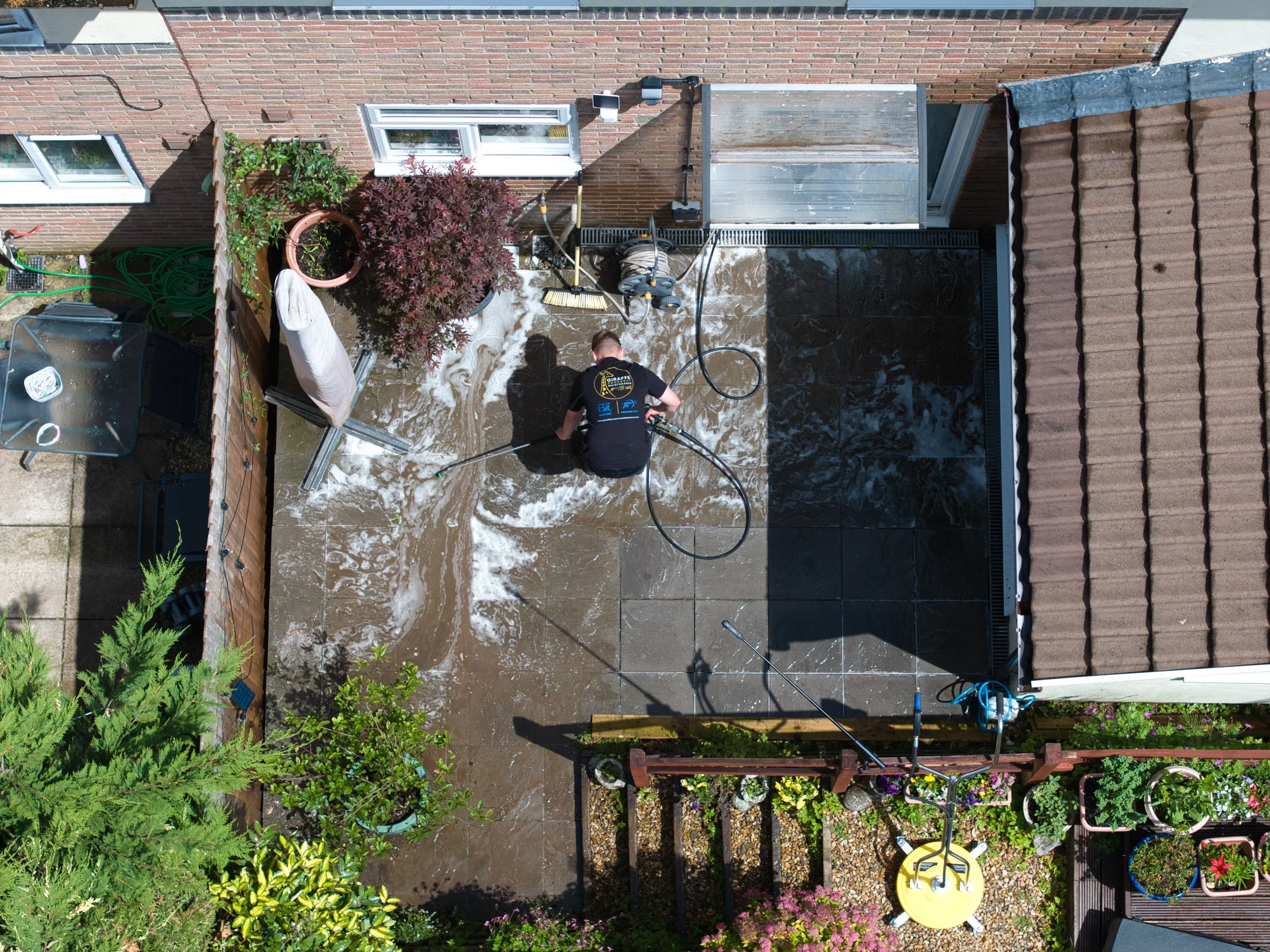 A person power washing a patio on a backyard terrace. The area features potted plants, a garden hose, and garden tools. The patio is adjacent to a brick house with windows and a sliding door.