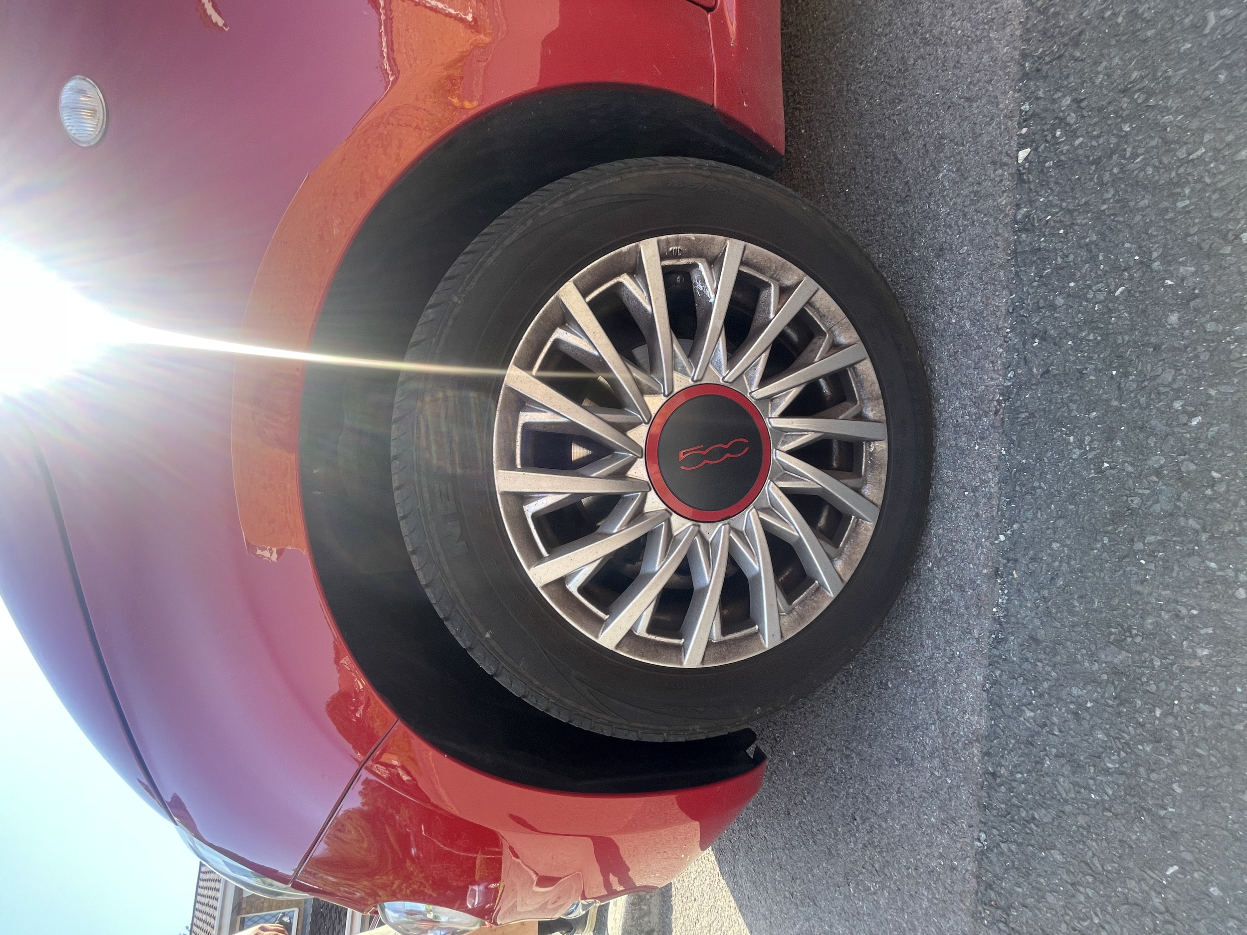 Red Fiat 500 car parked on a gravel lot, captured from a side angle, with the sun shining brightly, causing lens flare.