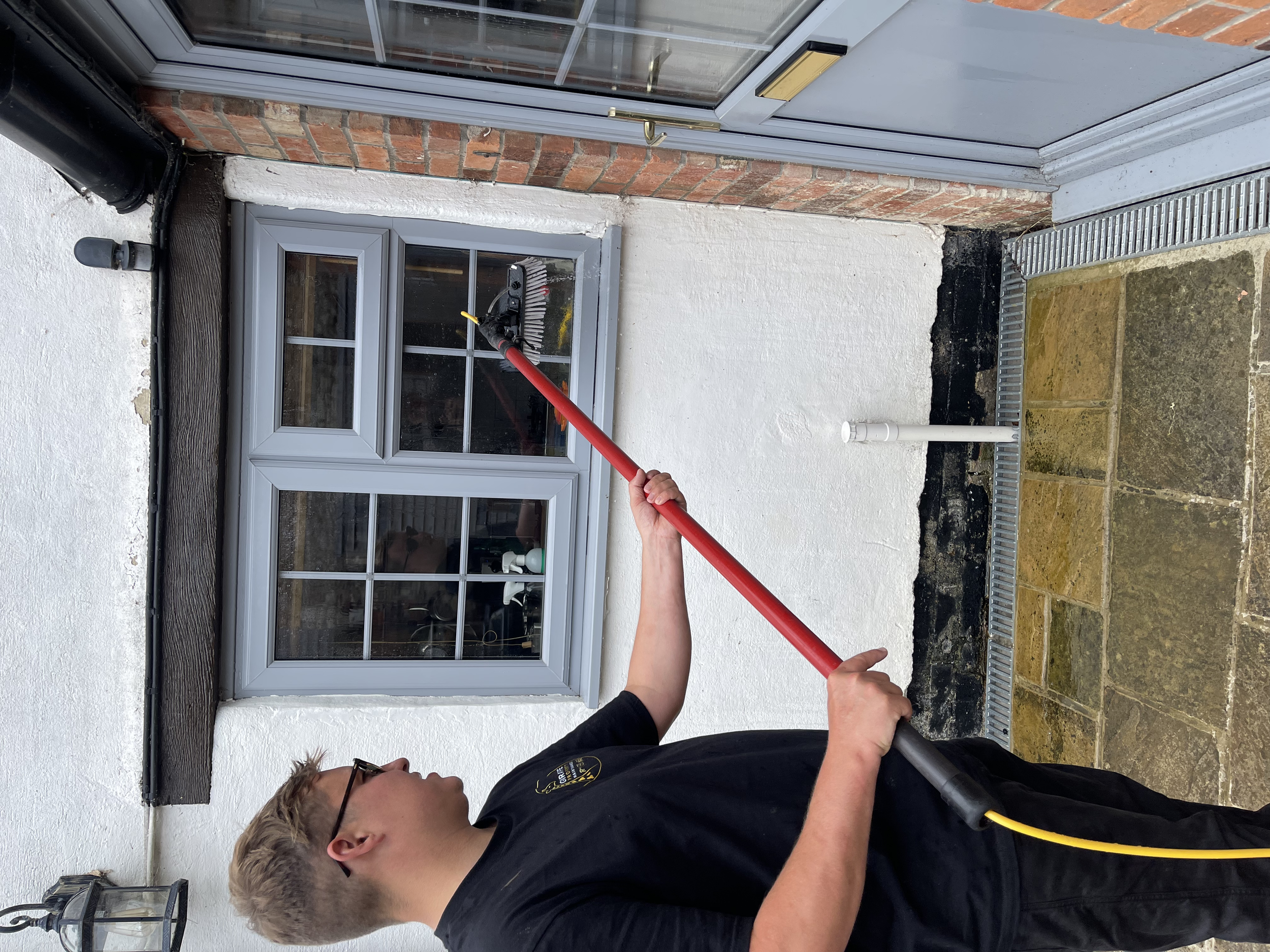 Person cleaning a window with a long-handled brush outside a house.