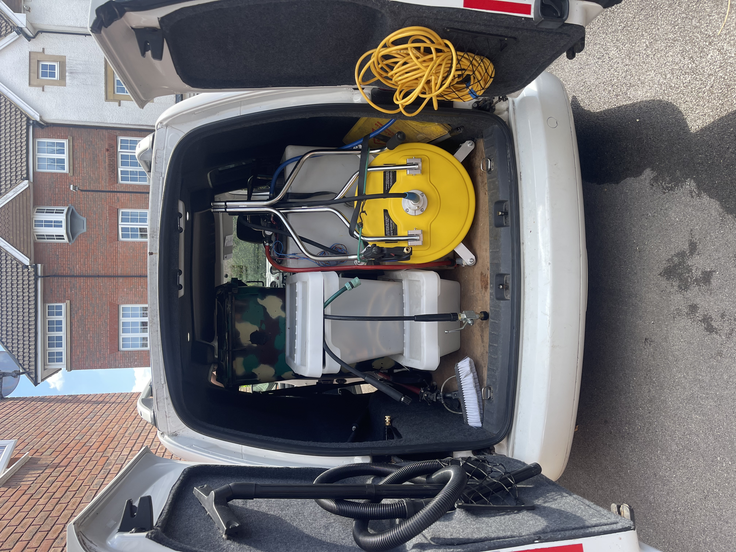 The open trunk of a white service vehicle revealing cleaning equipment including a yellow hose reel, a black vacuum hose, and various cleaning supplies. Residential buildings with brick and siding exteriors are visible in the background.