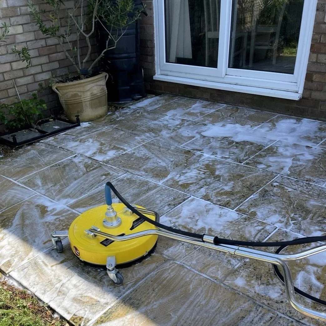 A pressure washer with yellow base cleaning a stone patio outside a house, with soap suds on the surface, near a sliding glass door, potted plants, and garden water cistern.