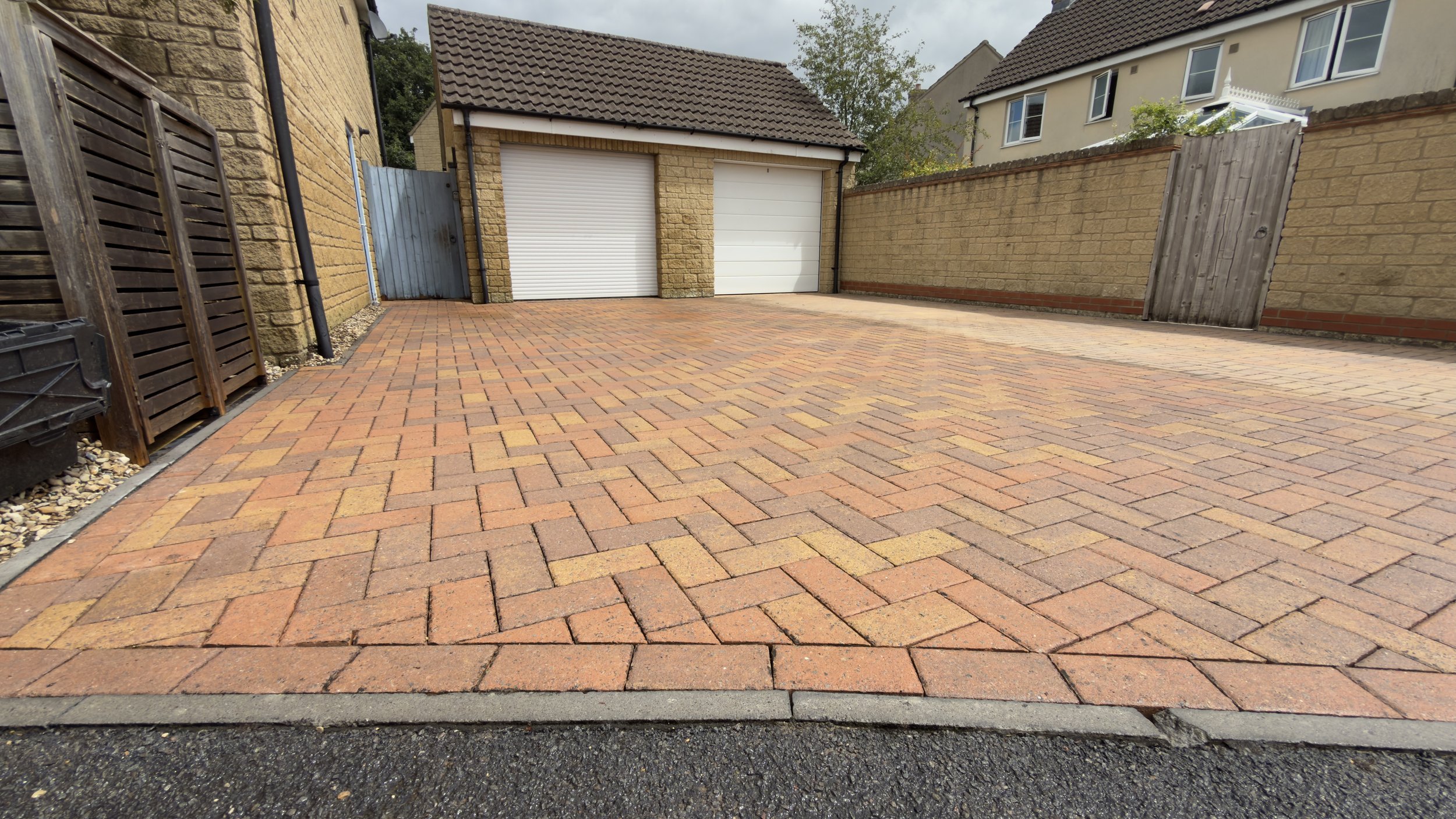 A paved driveway with orange and brown bricks, bordered by a black asphalt edge, next to a beige brick wall, with a small wooden gate and a garage with white roller doors. In the background, there is a house with a tiled roof and a window with white 