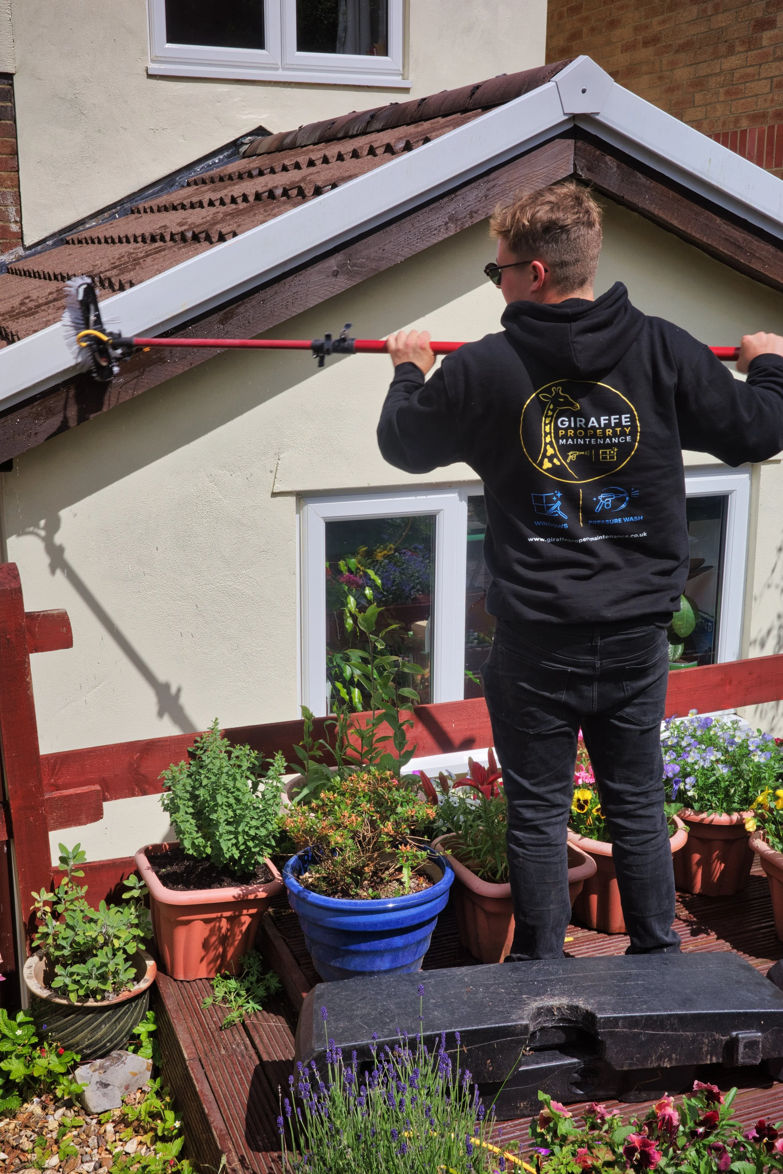 A young man wearing sunglasses and a black hoodie with a 'Giraffe Property Maintenance' logo is watering plants on a garden patio. There are potted plants and flowers on the wooden deck, and a house wall with a window in the background.