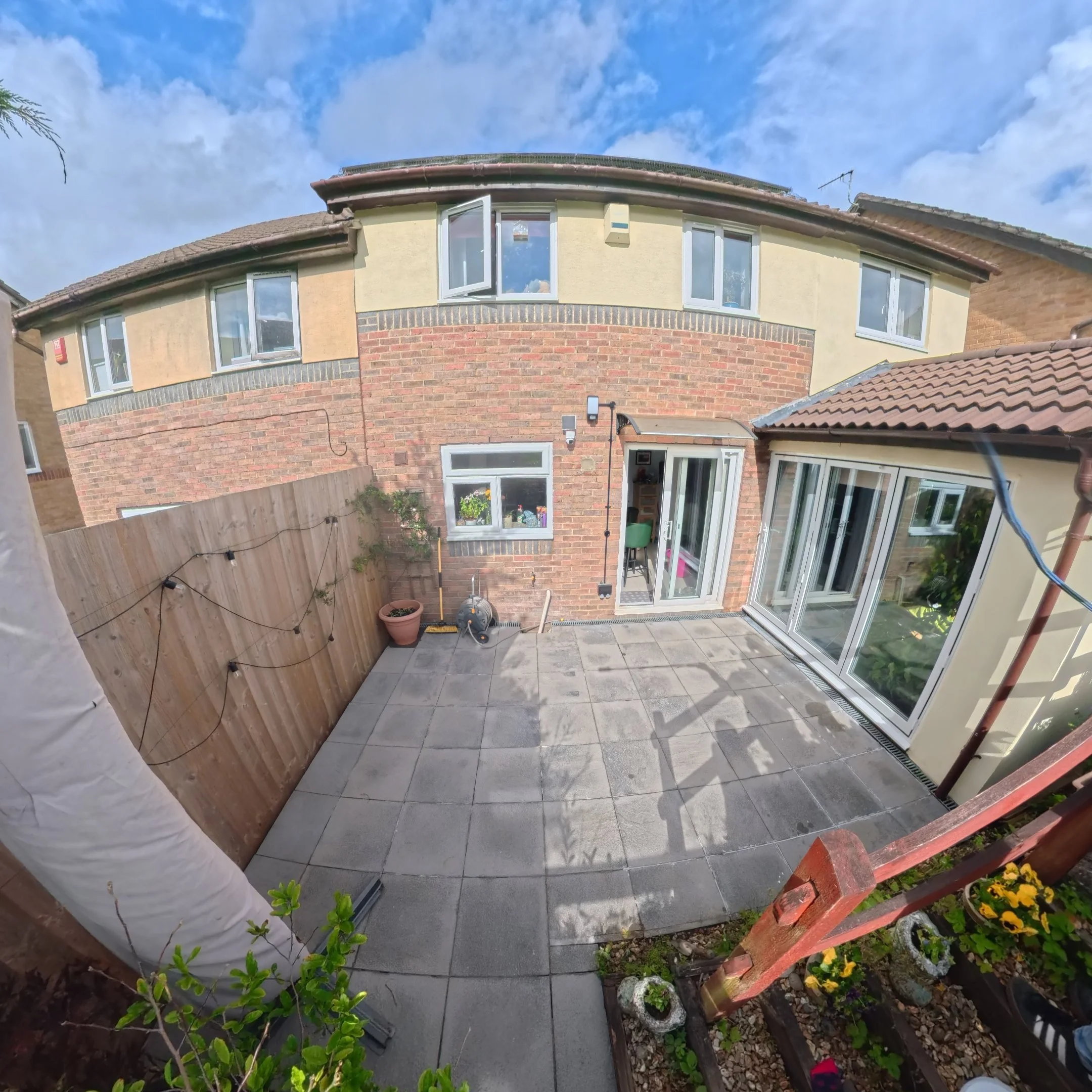 Backyard patio with grey tiled flooring, potted plants, a wooden fence, and a brick house with multiple windows under a partly cloudy sky.