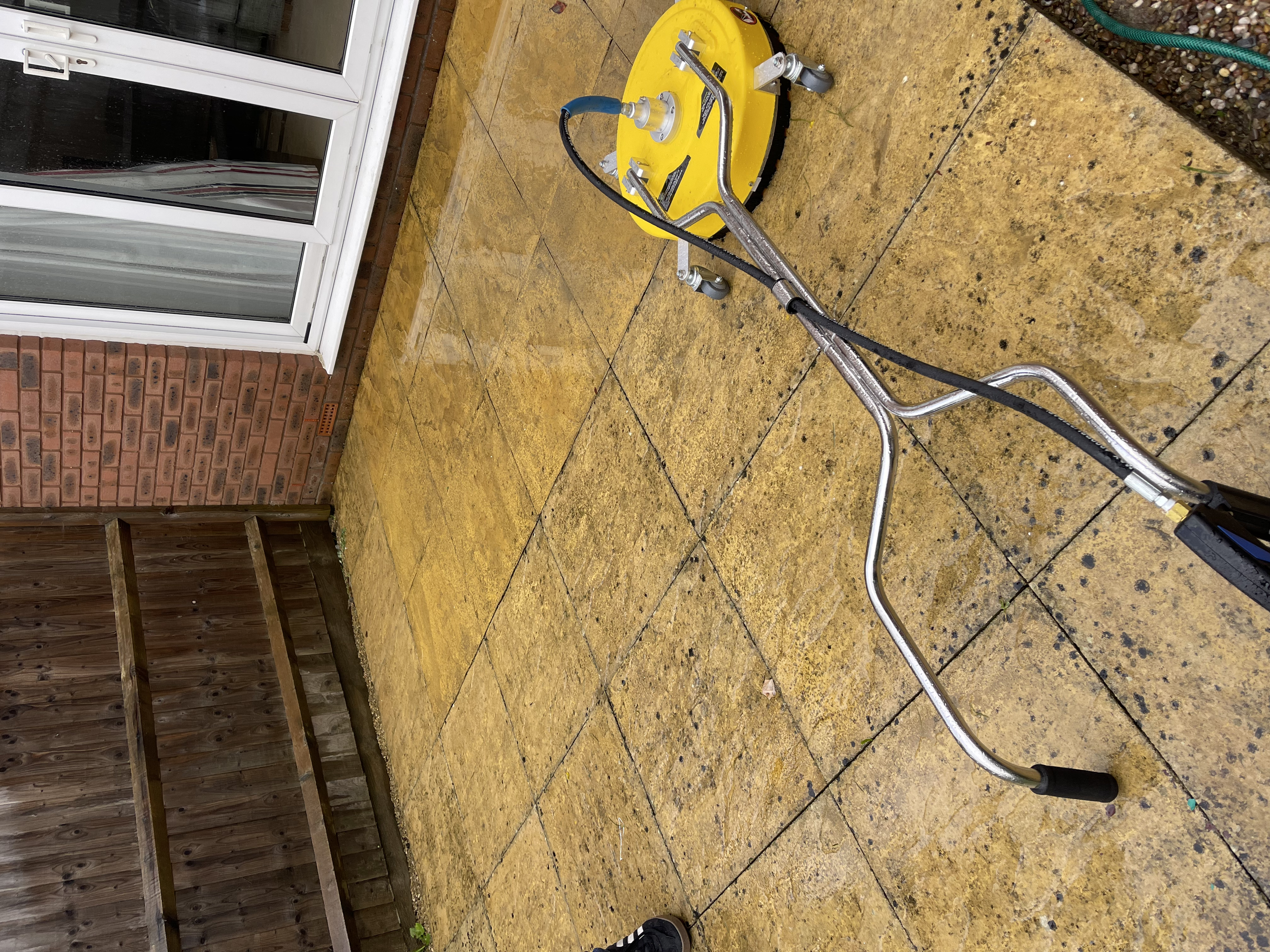Close-up of a yellow wet floor cleaning machine on a stone patio, with a wooden fence and brick wall in the background.