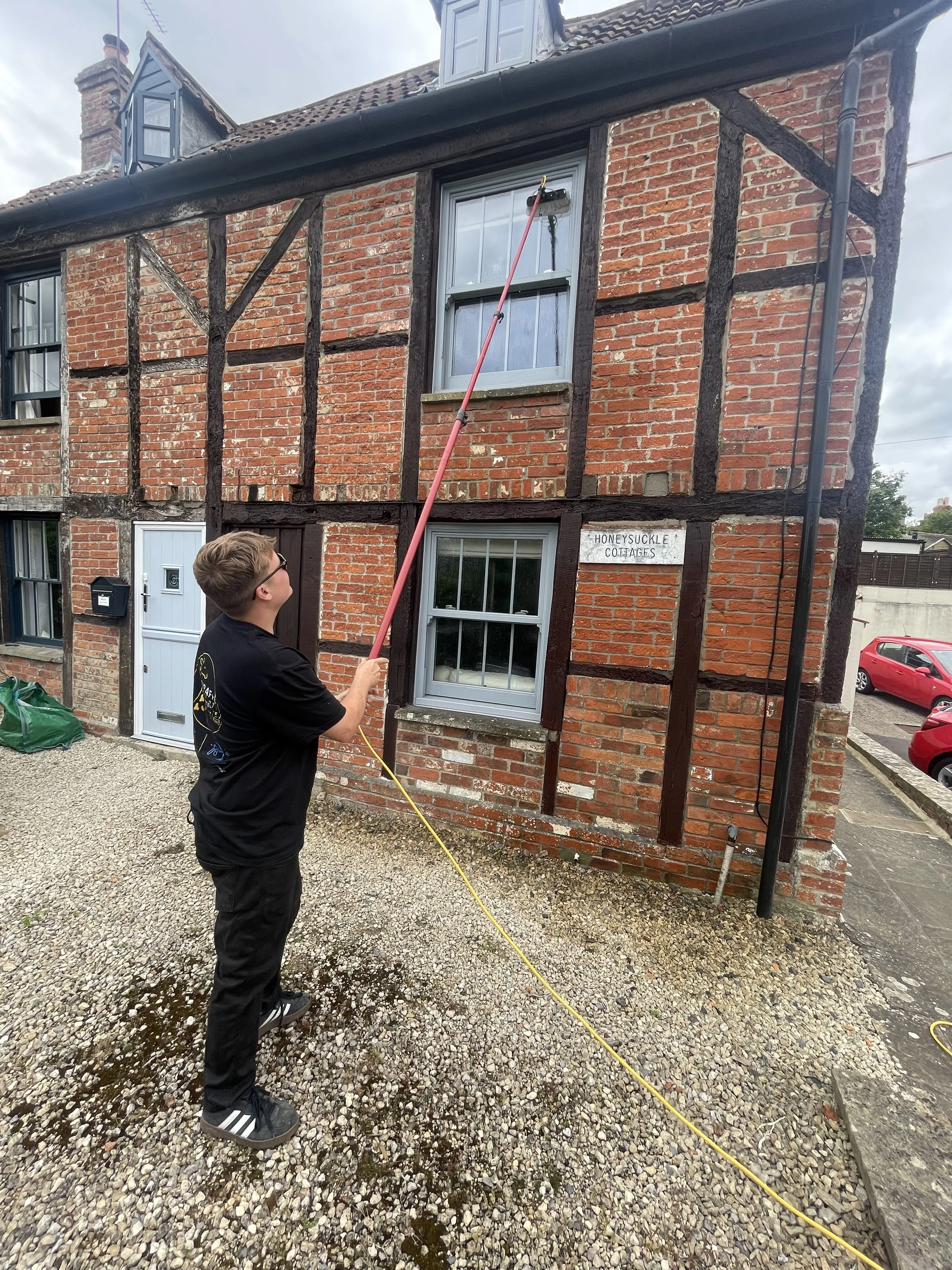 A young man in black clothing and glasses is cleaning the exterior of a brick house with a pressure washer that is creating water and foam on the gravel driveway. The house has timber framing, white-framed windows, and a sign that reads 'Honeysuckle Cottages'.