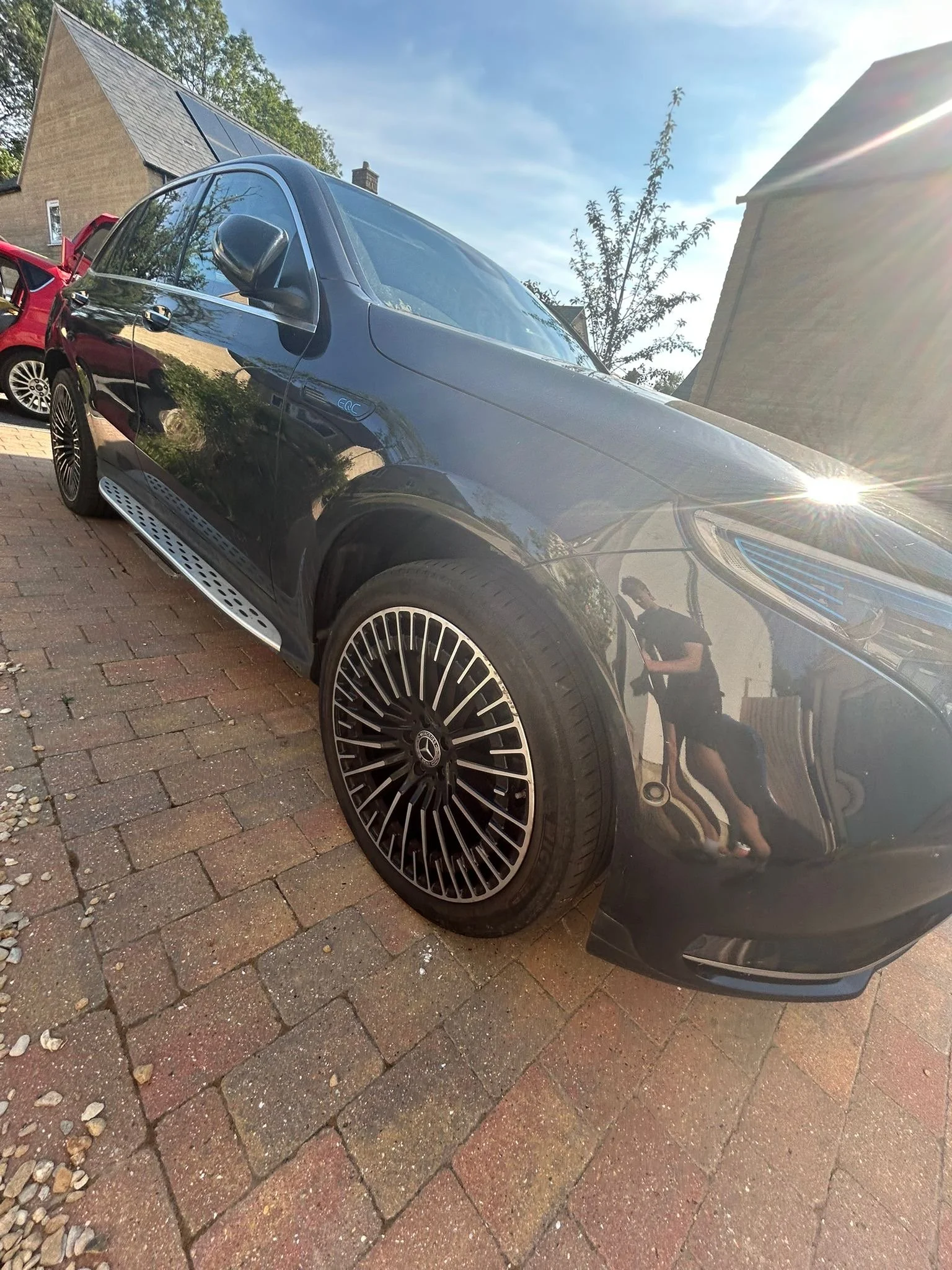 A black Mercedes-Benz parked on a brick driveway with a reflection of the person taking the photo visible on the car's surface. In the background, there are other cars, a house, trees, and a clear sky with the sun shining.