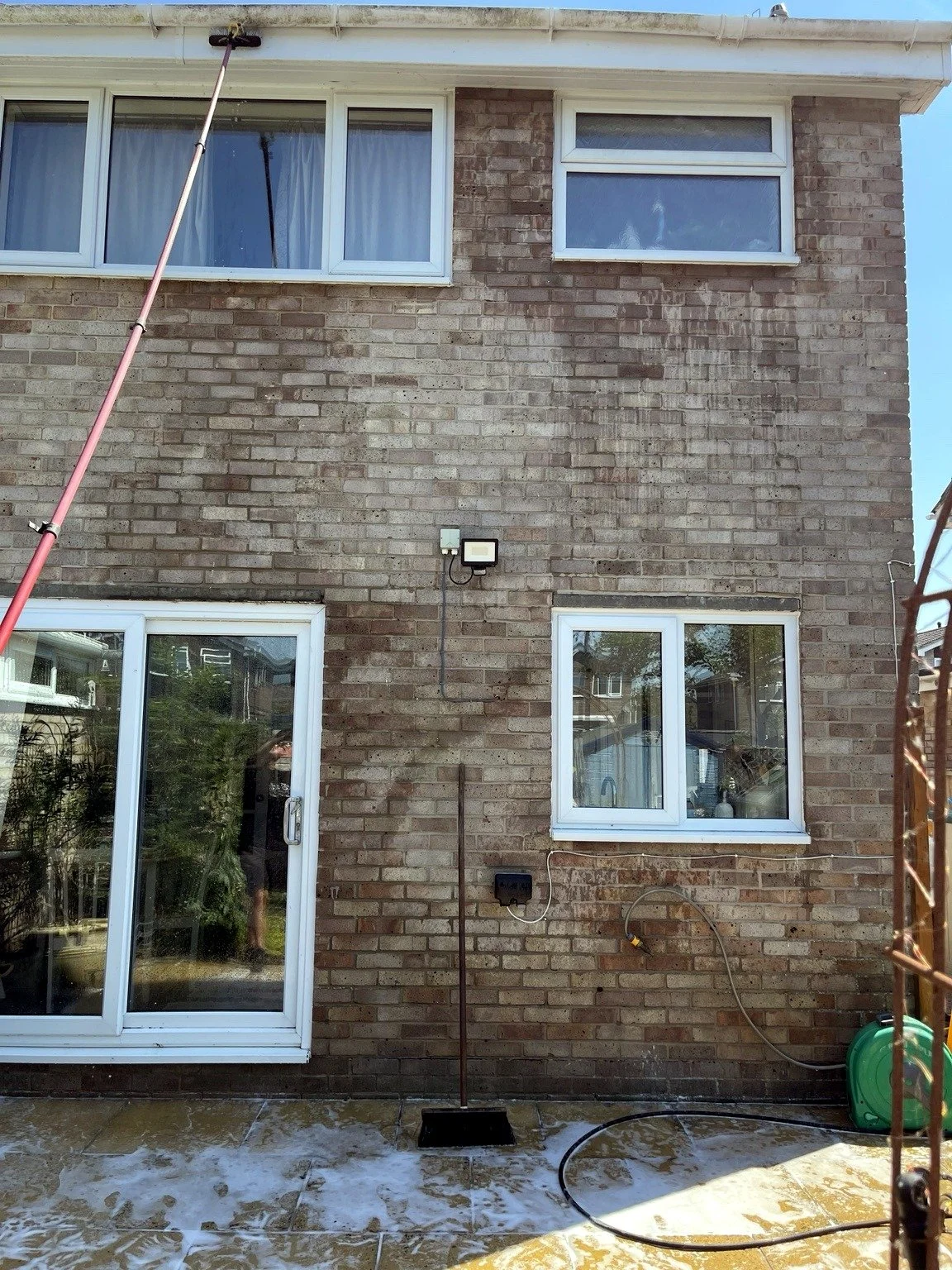 Back of a brick house with three white-framed windows; two on the upper level, one on the lower level, and a sliding glass door on the left. A pole extends from the ground to the roof on the left side, and there is a black stake and a garden hose ree