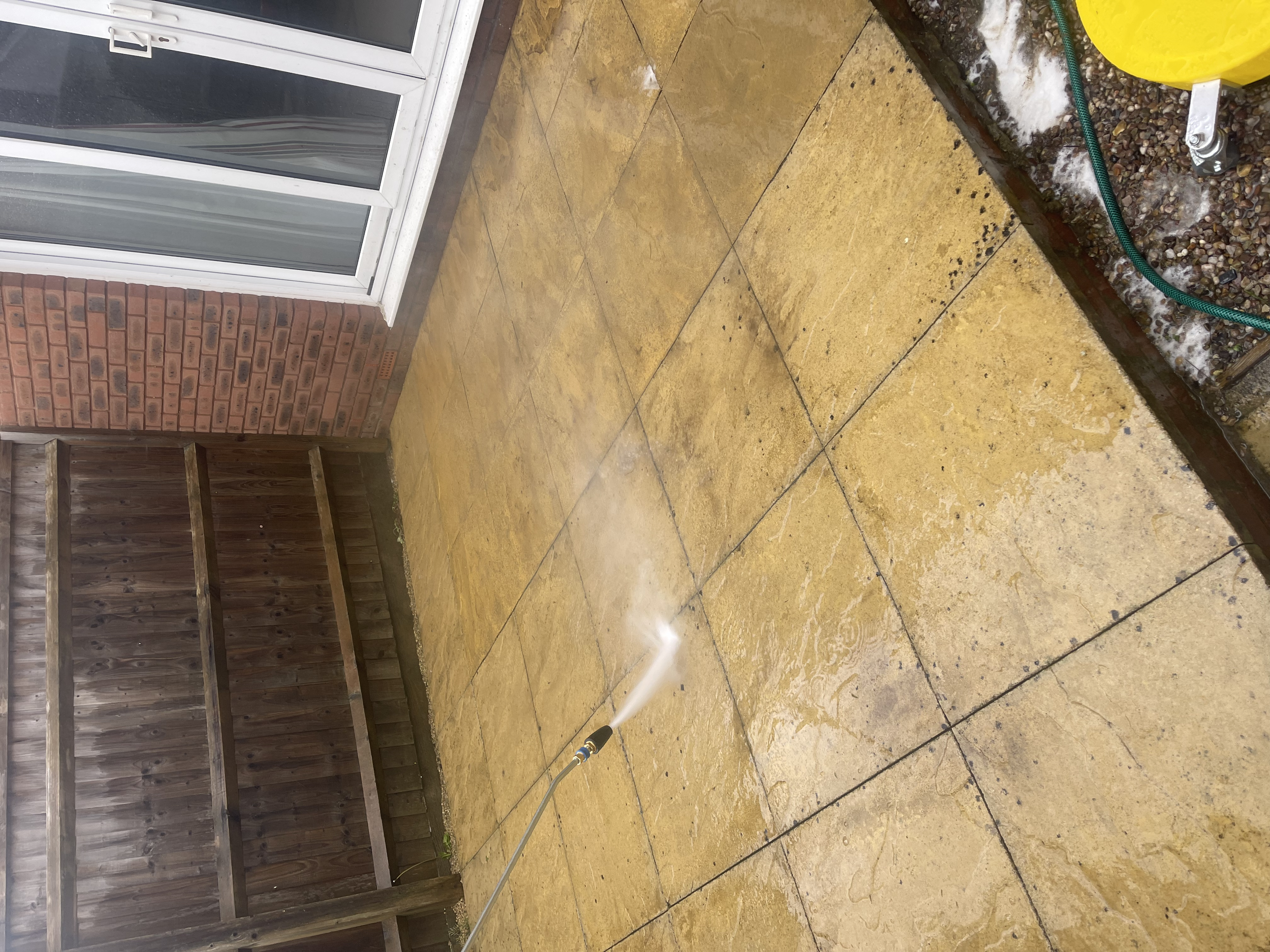 A garden patio area with wet yellowish tiles, a red brick wall, a wooden fence, and a yellow water container connected to a green hose, with water spray on the tiles.